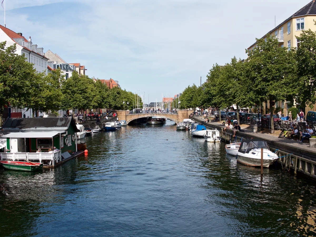 A tree-lined Christianshavn canal in Copenhagen under a bright blue sky with houseboats and moored boats.