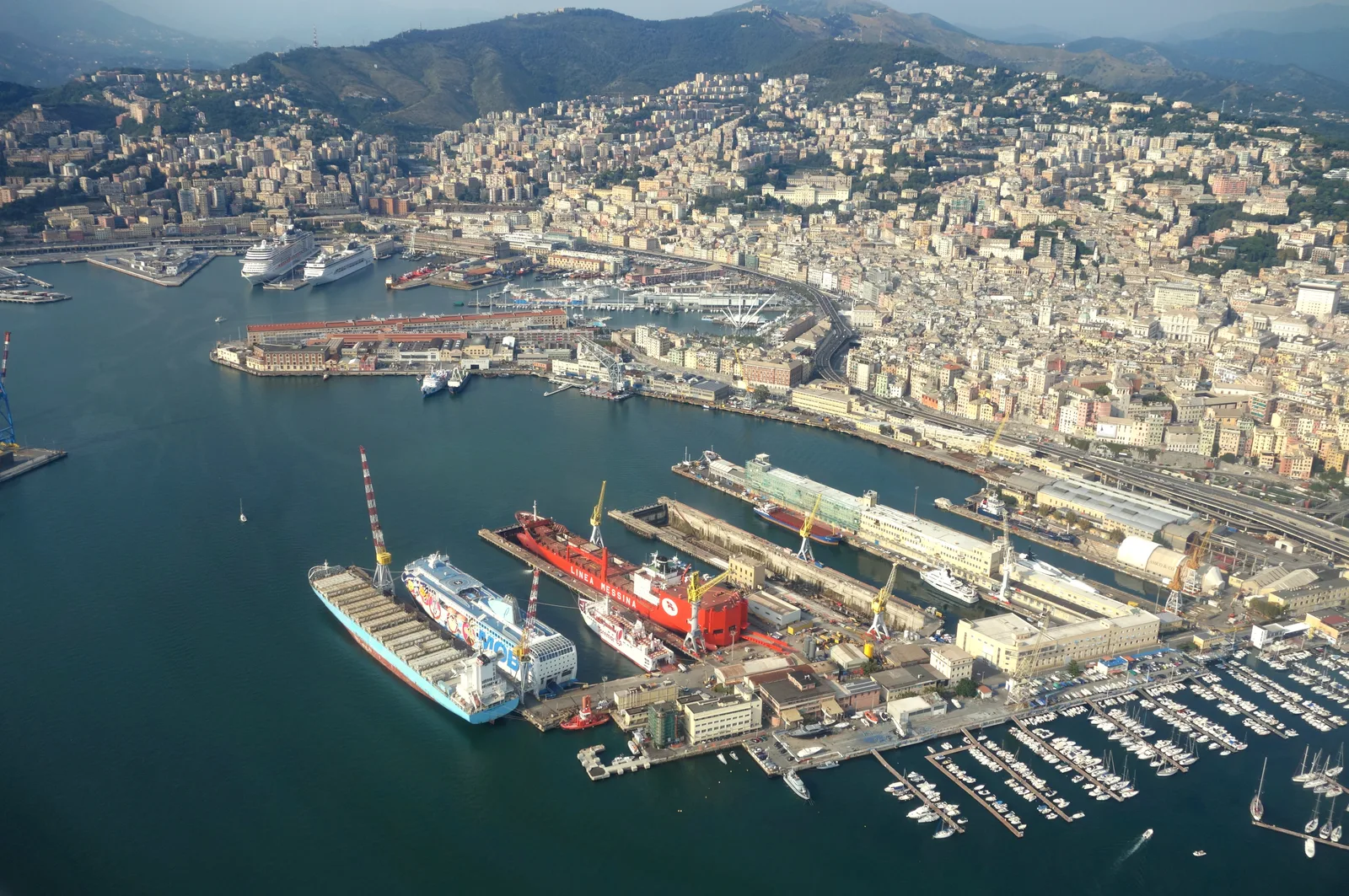 An aerial view of Genoa's harbour with cruise and cargo ships, docks and the city terraced up the hills.