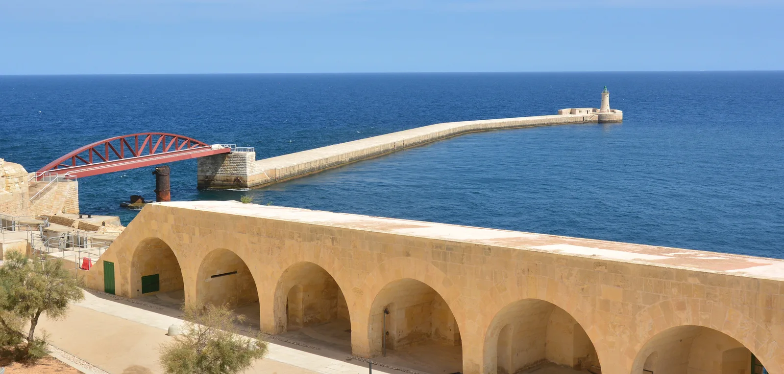 A long stone breakwater with a red bridge and lighthouse at the entrance to Grand Harbour under bright blue sky.