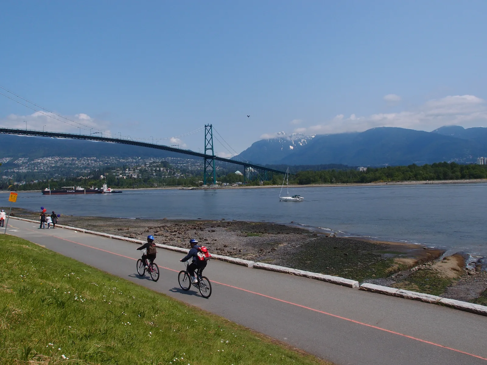A seawall path at Stanley Park with the Lions Gate Bridge, boats and mountains across the water and two cyclists in the distance.