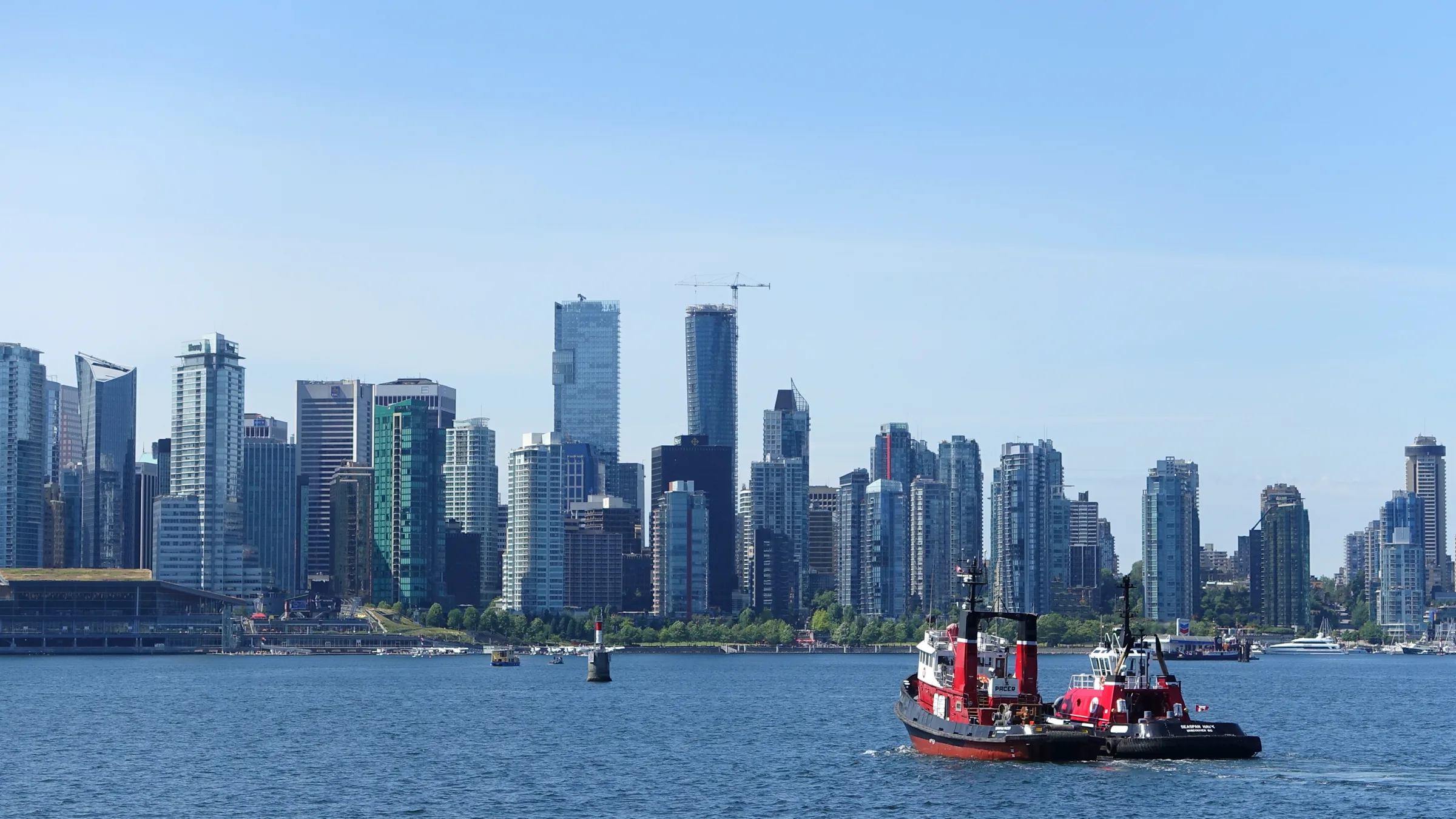 Downtown Vancouver towers rising above Burrard Inlet with two red tugboats on the water under a clear blue sky.