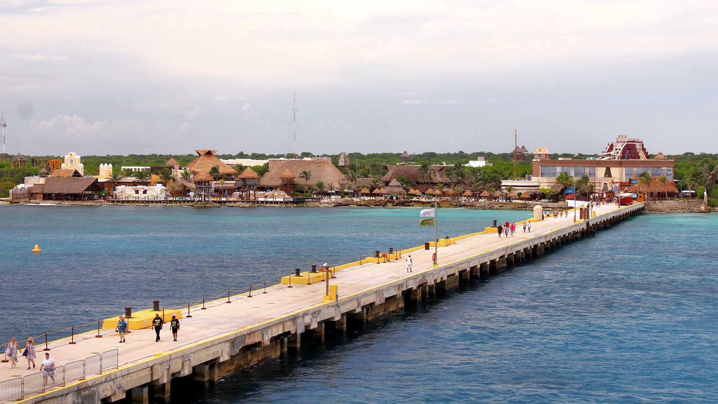 The long cruise pier of Costa Maya reaching towards the thatched port village with a few visitors walking along it.