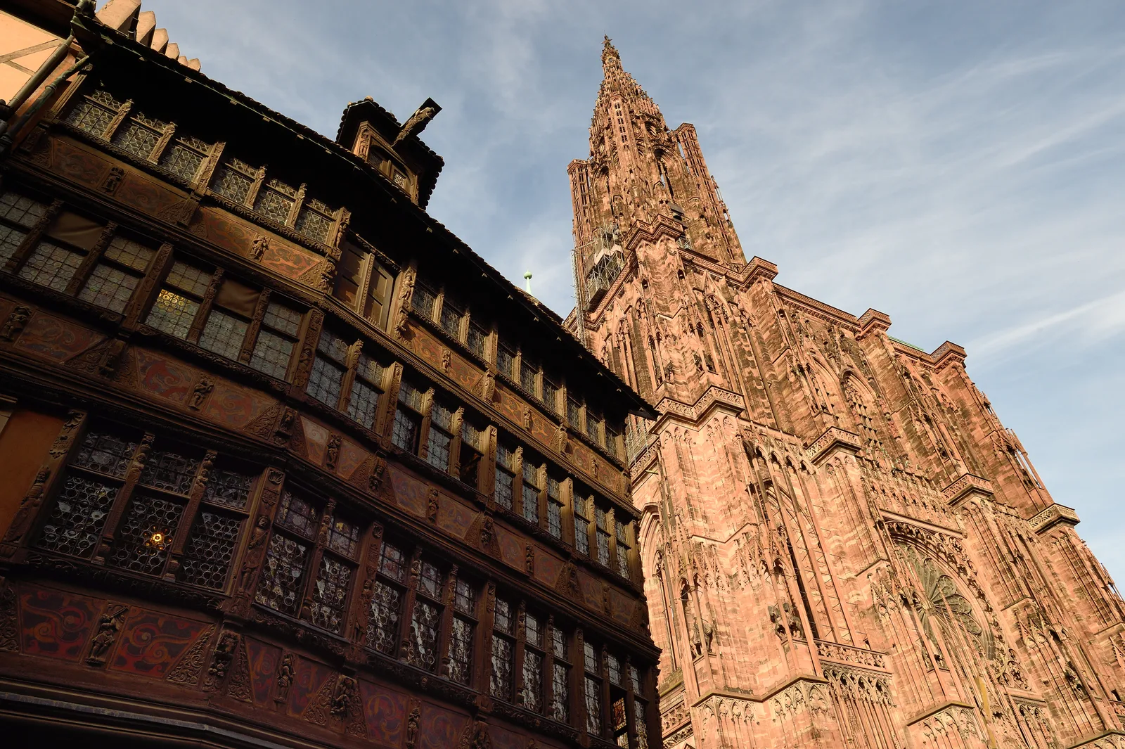 The carved wooden facade of Maison Kammerzell beside the pink sandstone tower of Strasbourg Cathedral against a blue sky.