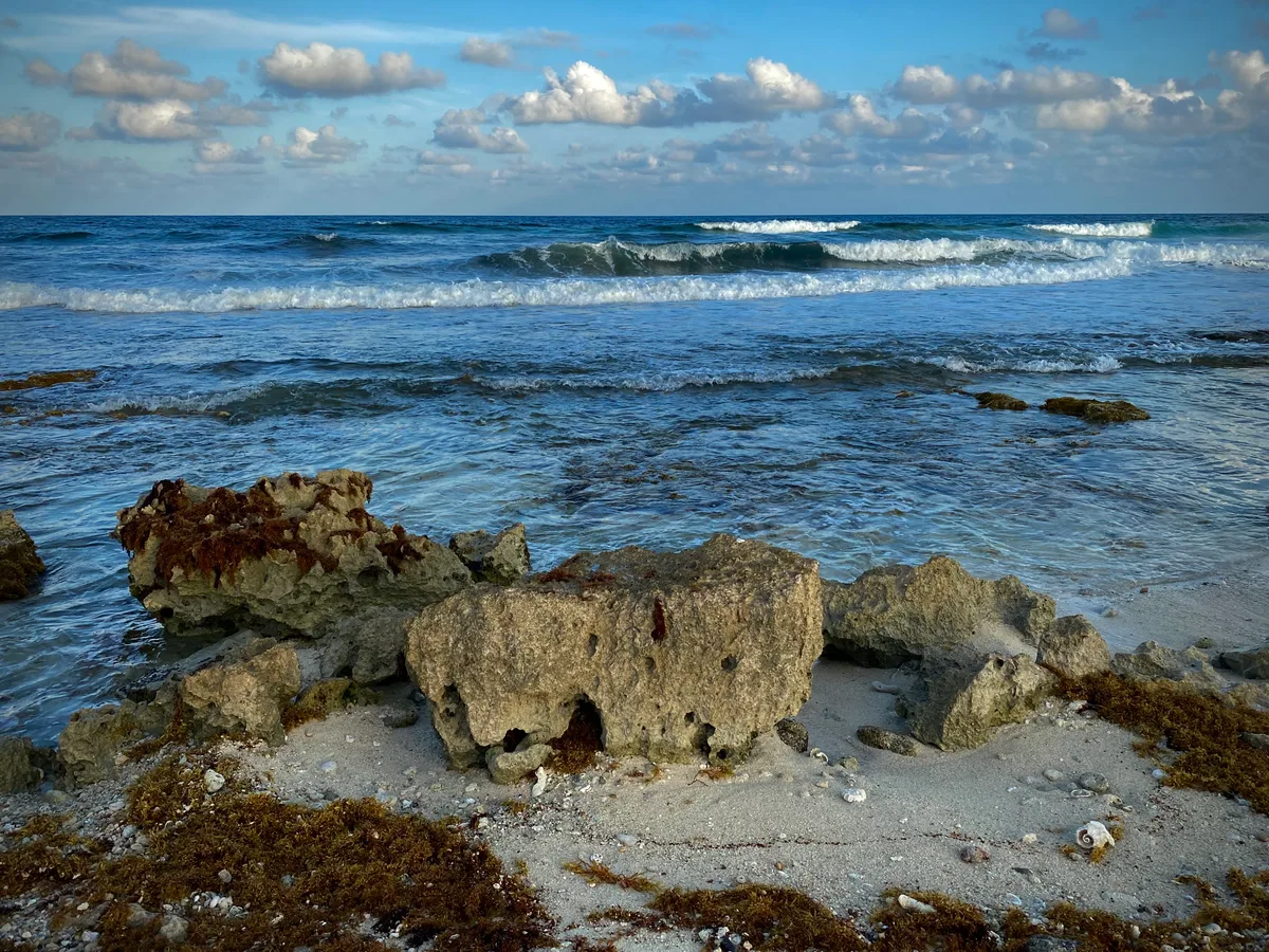 Waves break over rough rocks and sand on Cozumel's wild eastern coast under scattered clouds.