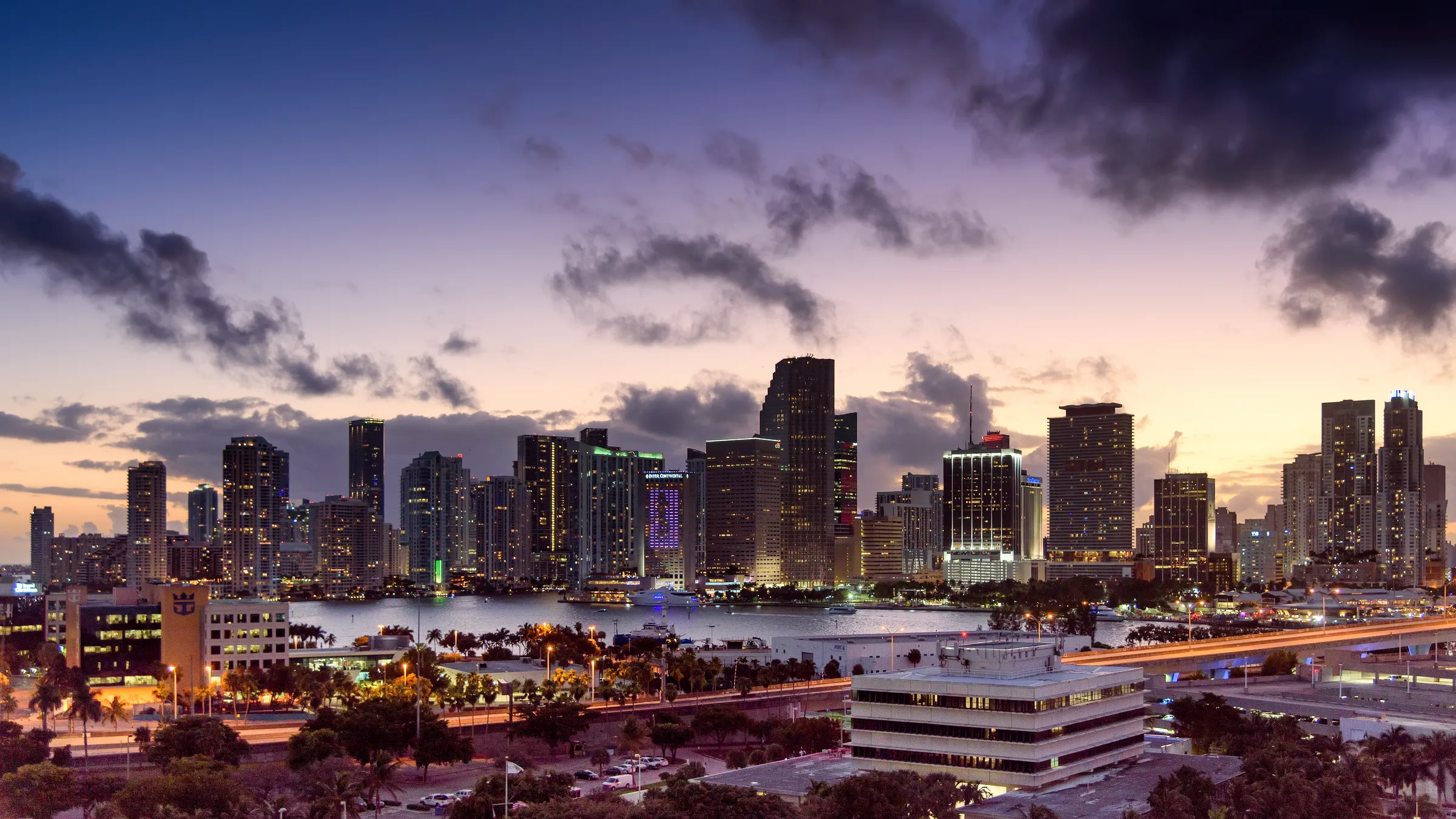 Evening skyline of downtown Miami with lit towers across Biscayne Bay under a purple and orange sunset sky.