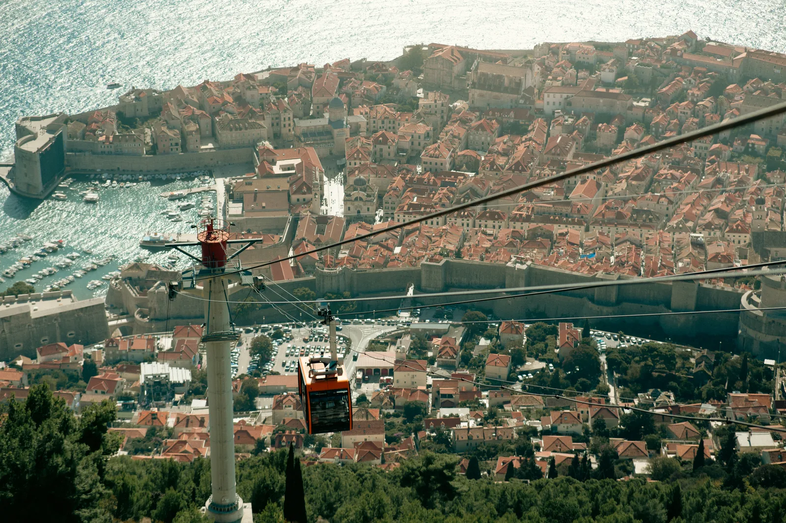 A red Dubrovnik cable car beside a tall pylon above the orange rooftops and walls of the old town and the harbour.