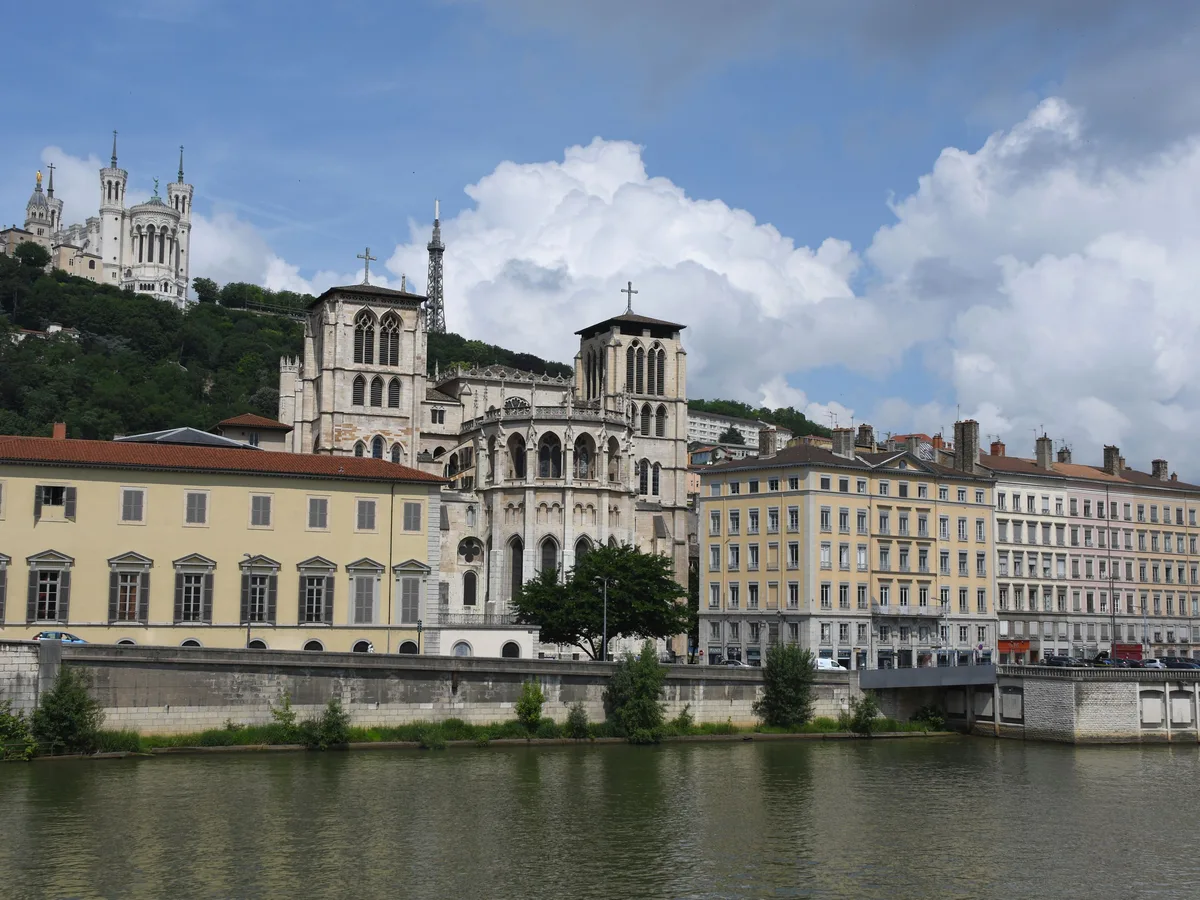 The Saône riverbank at Lyon with the cathedral of Saint-Jean-Baptiste, a long classical building and a church on the hilltop, on a bright day with clouds.