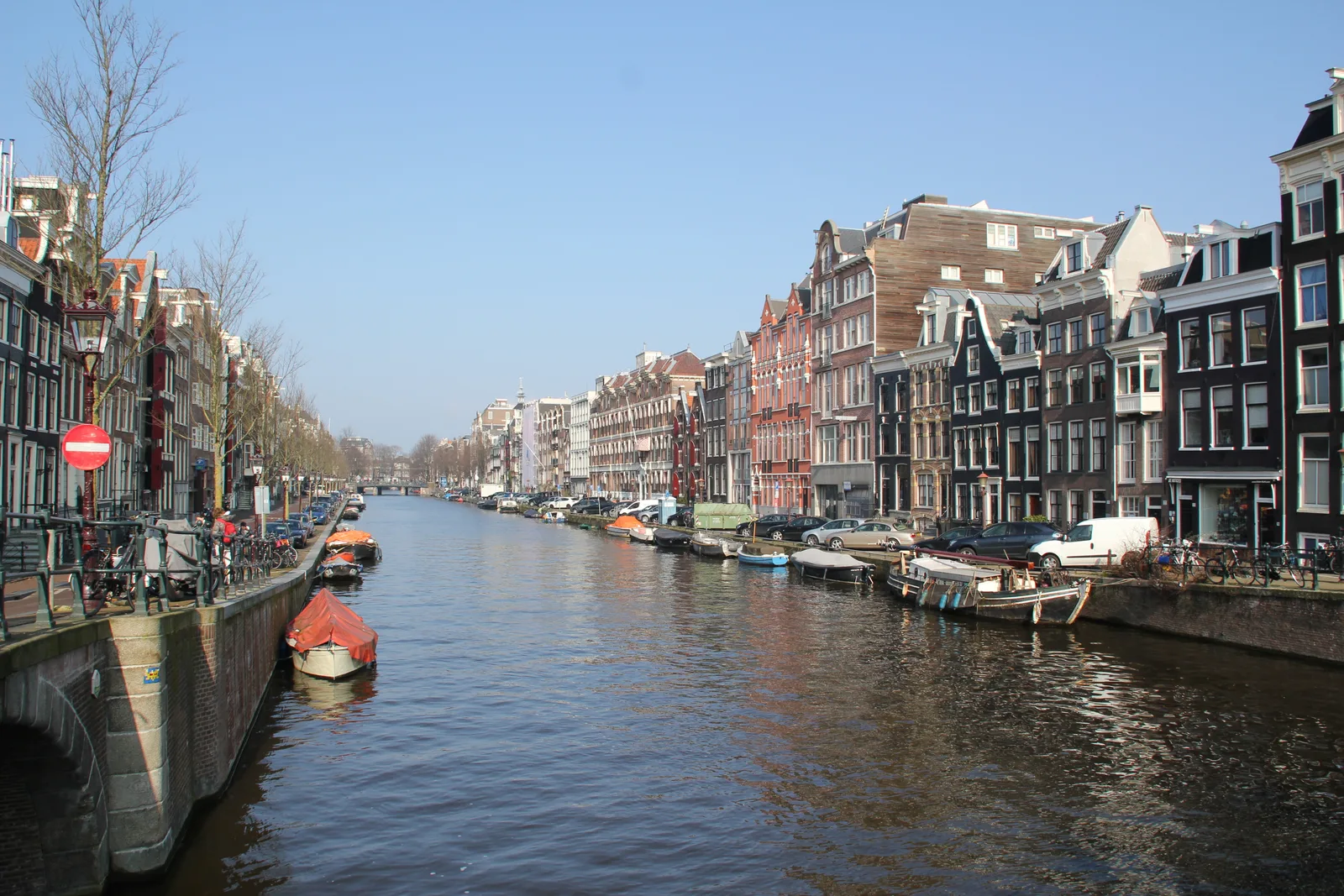 A canal in central Amsterdam lined with tall brick houses, moored boats and a bridge, under a bright blue sky.