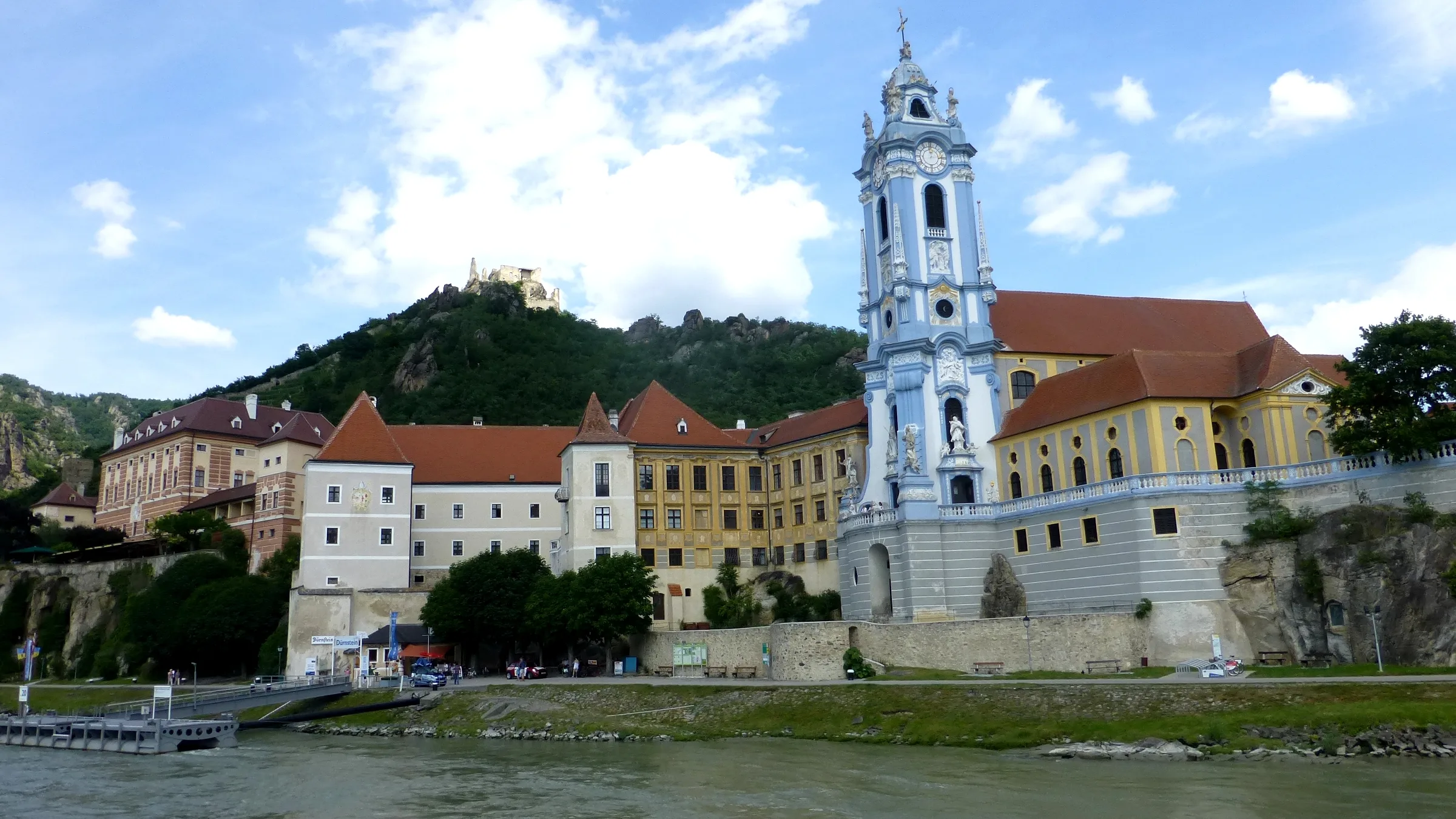 The abbey church of Dürnstein with its blue tower and the abbey wing beside the Danube, with the castle ruin on the ridge behind.