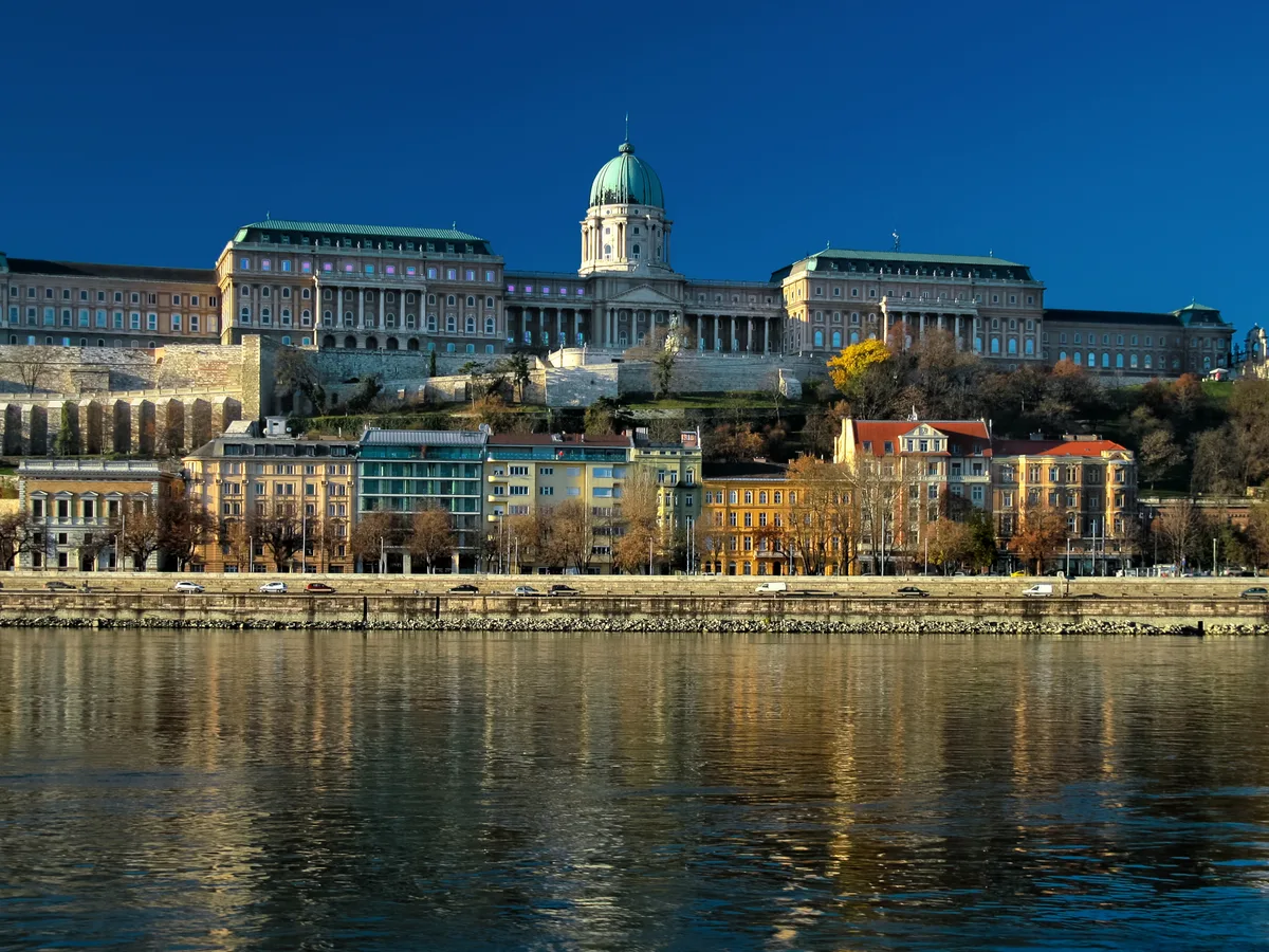 Buda Castle palace above the Danube embankment with autumn trees and the river reflecting the hillside.