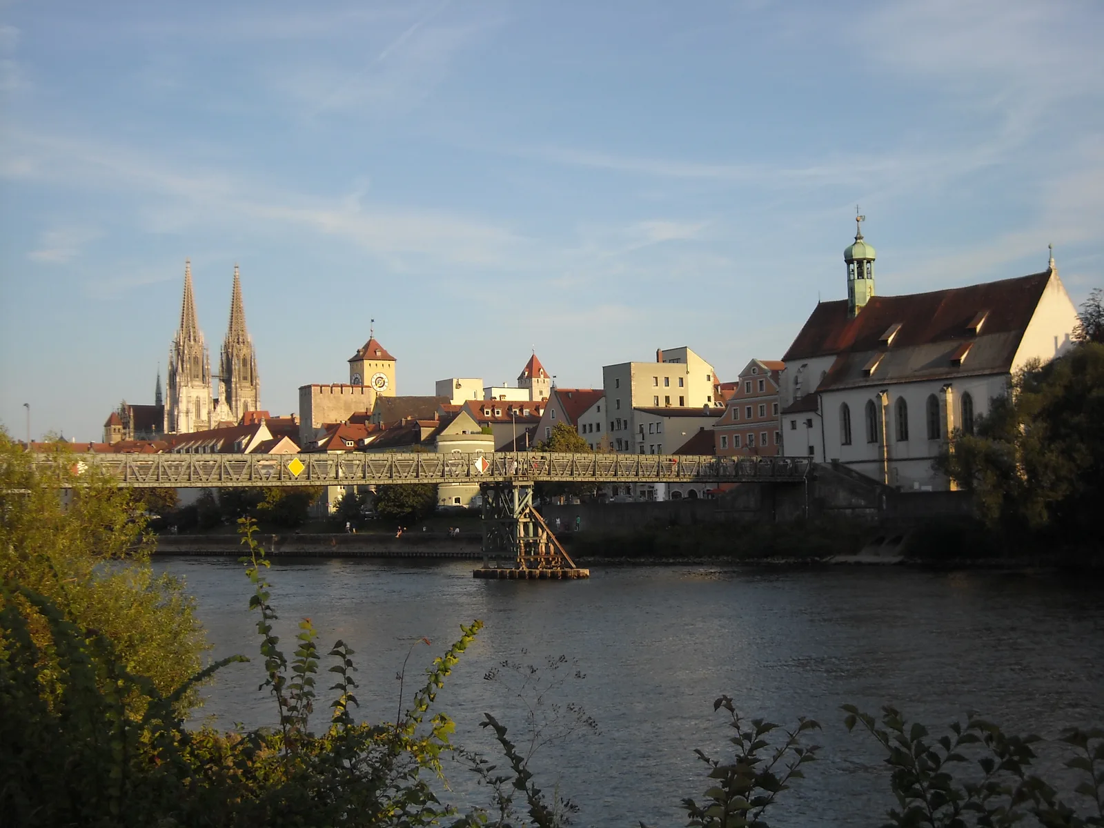 Regensburg old town seen from the north-west across the Danube, with the Iron Footbridge and two church spires in warm evening light.