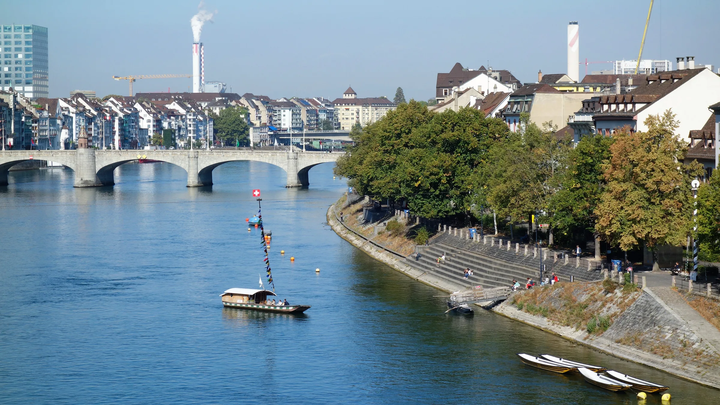 The Rhine in Basel with a bridge in the distance and a small ferry crossing under a clear blue sky.