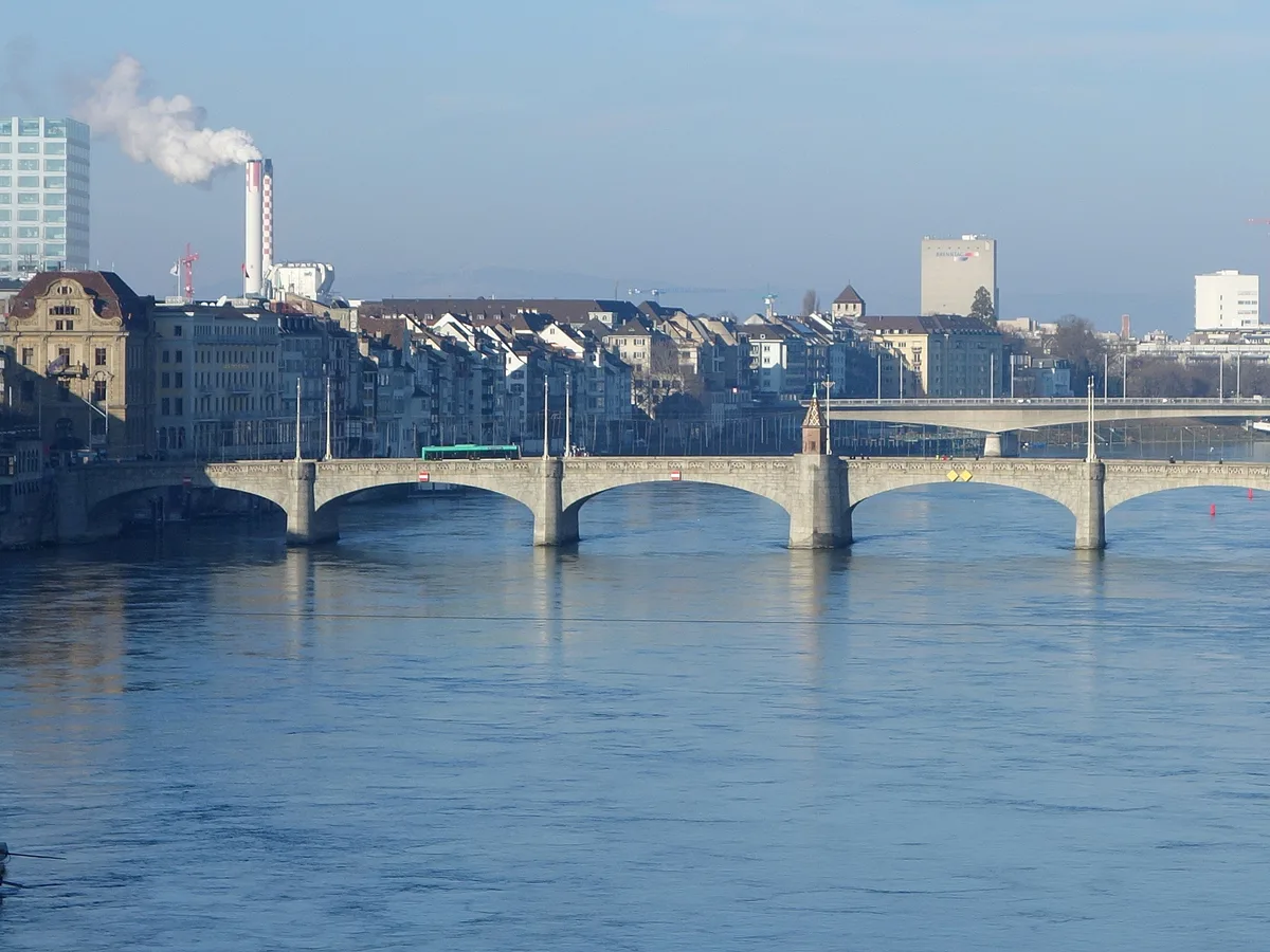 A wide view of the Rhine and the Mittlere Brücke arches in Basel with houses along both banks on a calm blue day.