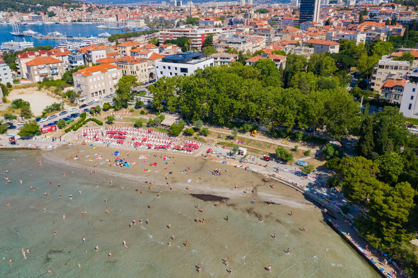 Aerial view of the curve of Bacvice beach with red parasols, the park and the rooftops of Split behind.