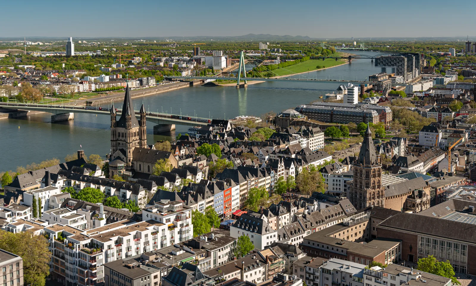 Aerial view over Cologne showing old-town roofs, the Rhine and a bridge in the distance.