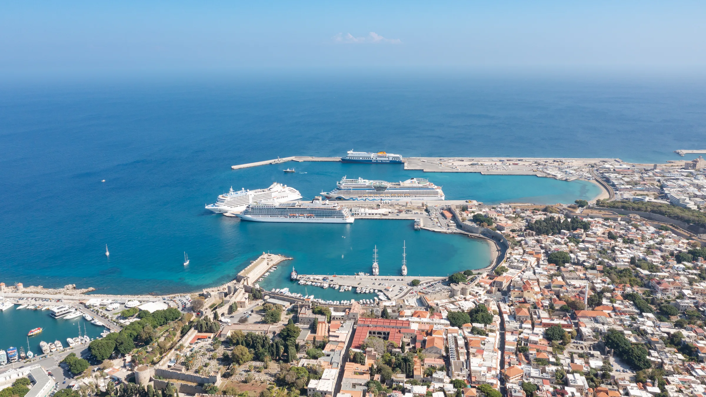 Aerial view over Rhodes Town with cruise ships docked in the commercial harbour, the old town's roofs and the coastline.