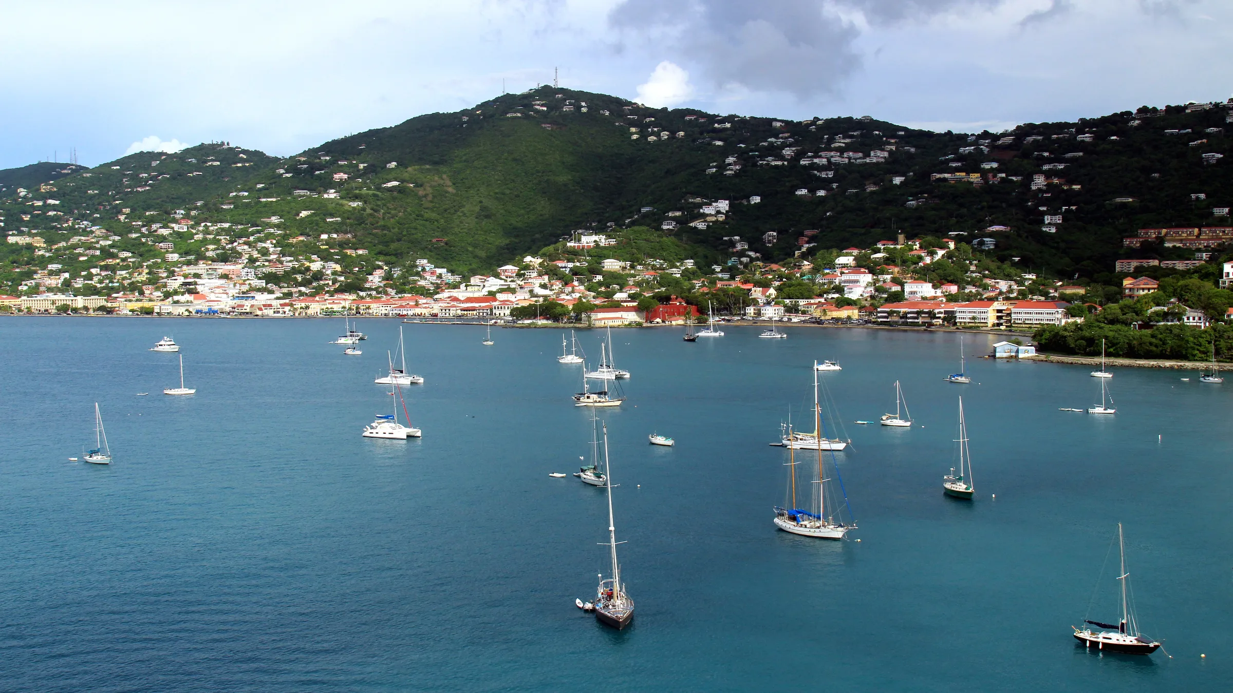 View over Charlotte Amalie harbour from a hillside with sailboats anchored in blue water and houses climbing the green hills.