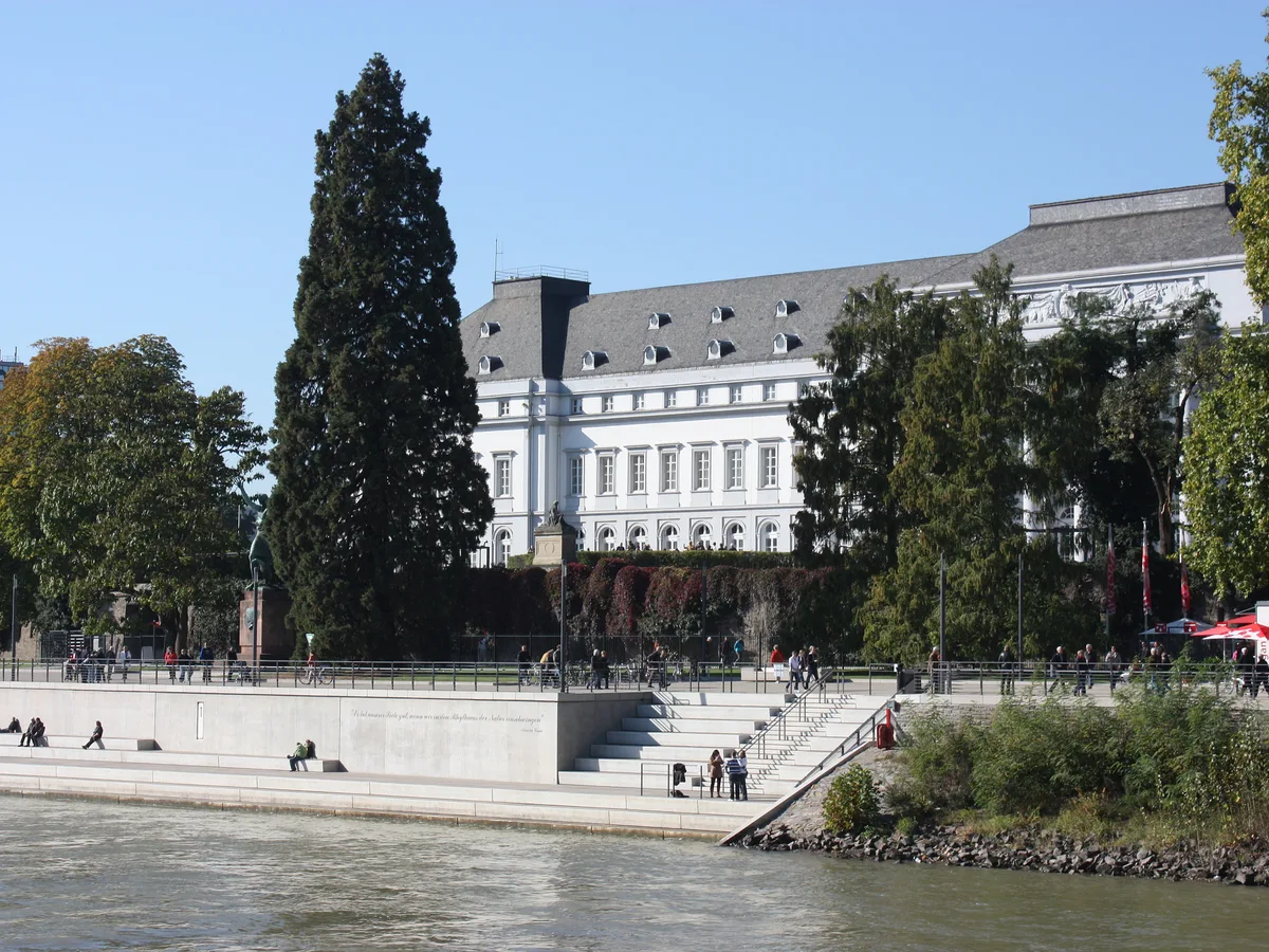 The cream facade of the Electoral Palace at Koblenz above a paved Rhine promenade with steps and a few distant visitors.