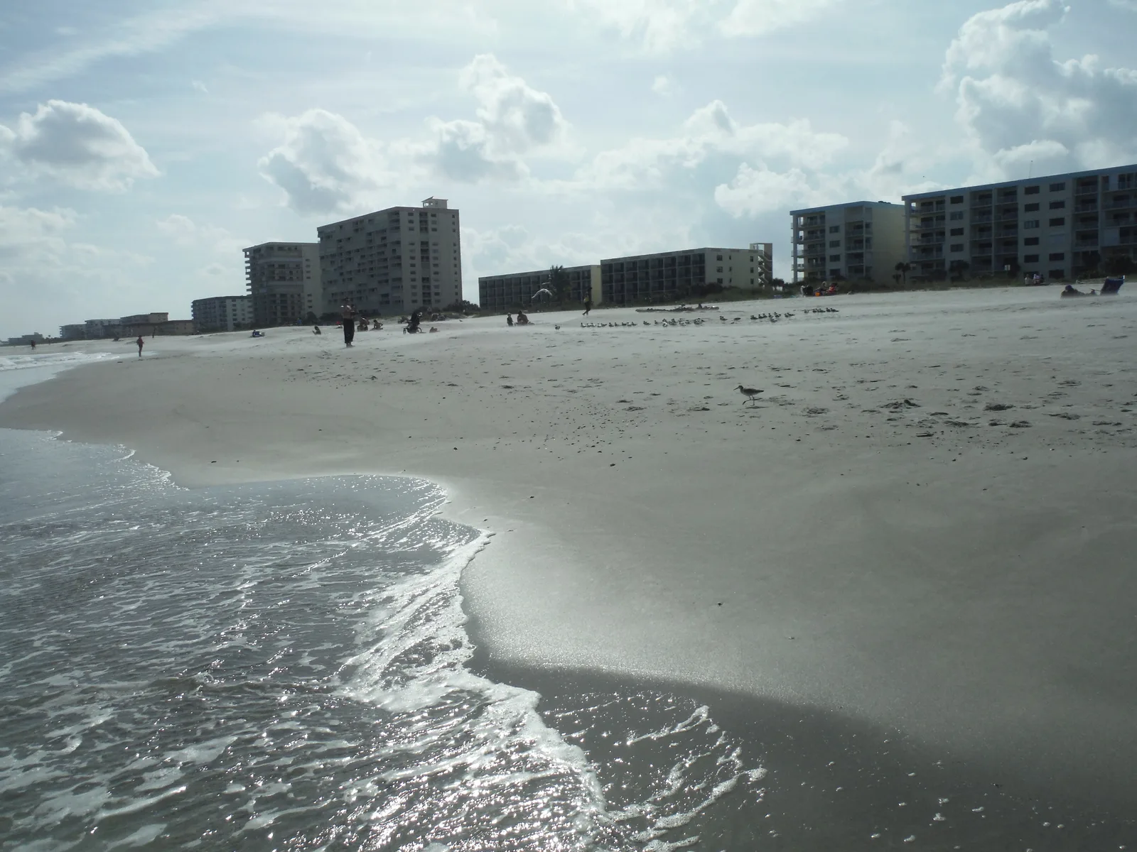 Gentle waves washing onto a gray-sand beach with low beachfront buildings in the distance at Cocoa Beach.