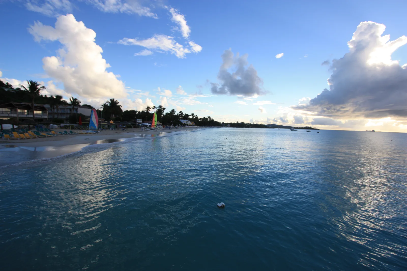 A calm, curved beach with a line of palms and small sailboats in the distance on Dickenson Bay, Antigua.