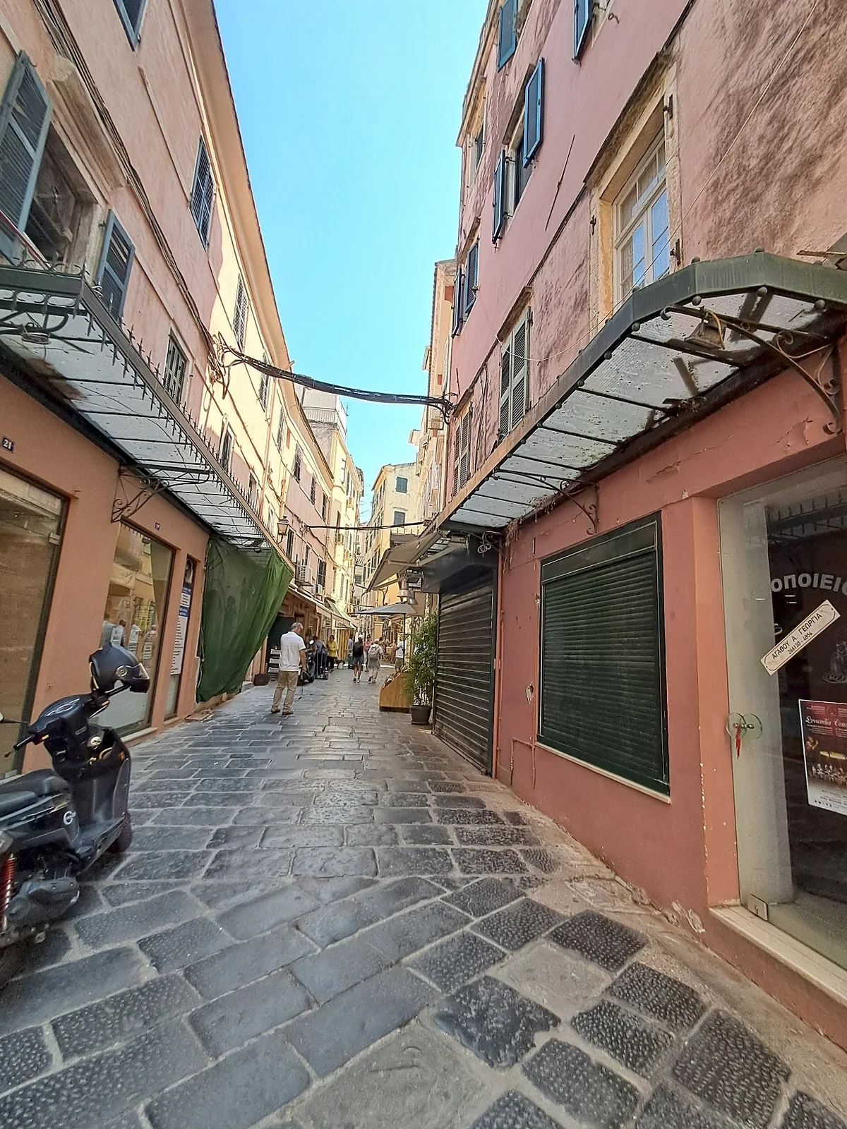 A narrow cobbled Corfu Old Town street between pink buildings with shop awnings and a scooter parked on the left.