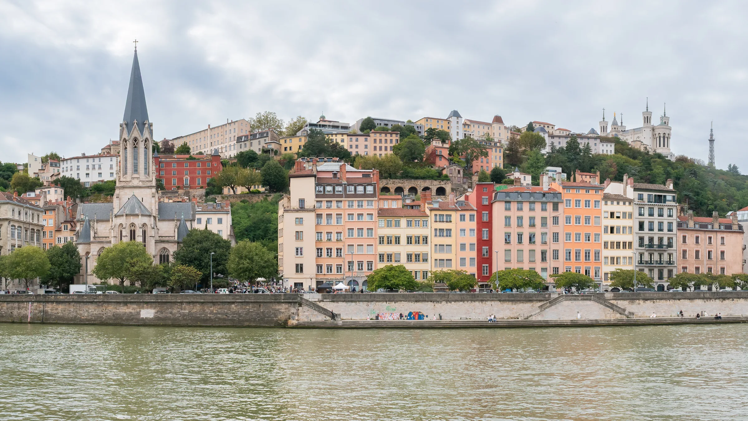A very wide view of the Quai Fulchiron in Lyon with pastel houses and a church spire along the Saône under a wooded hill.