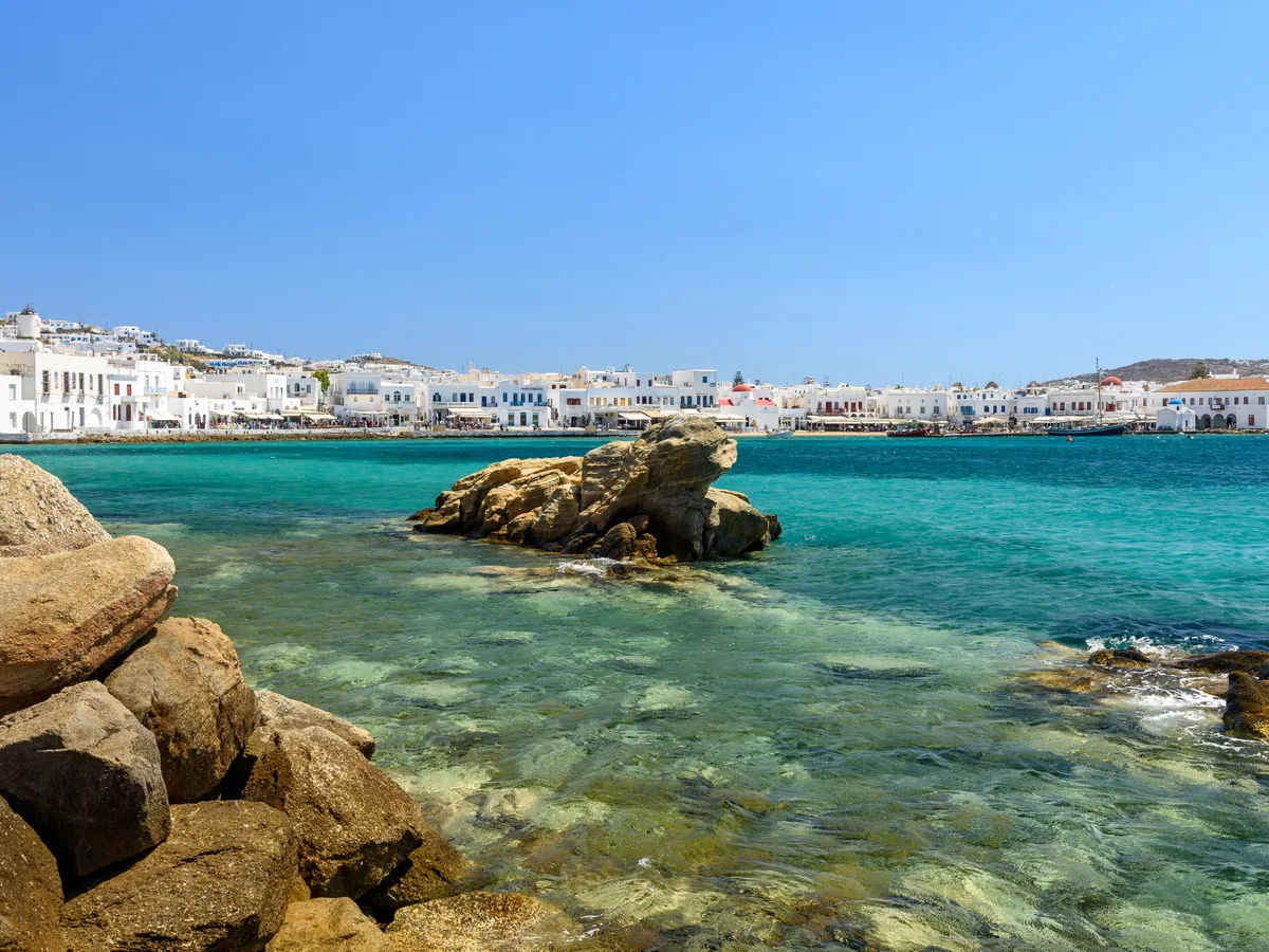 Turquoise water and rounded boulders in the foreground with the Mykonos waterfront stretching along the far shore.