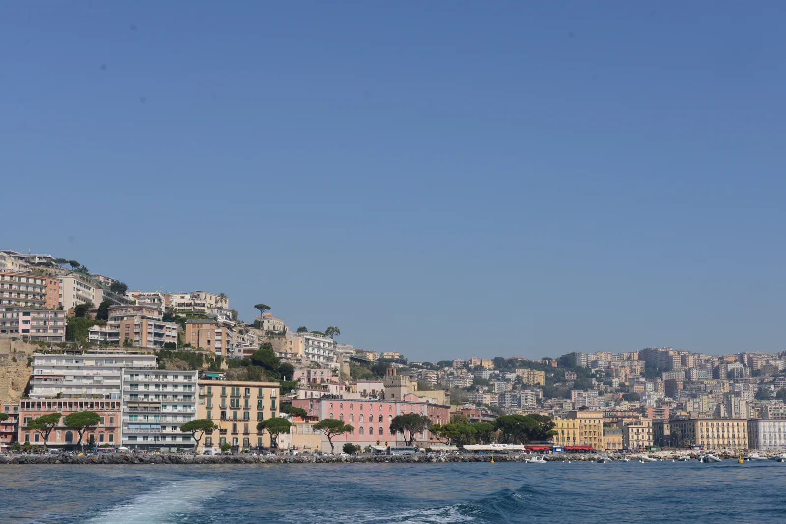 Naples seen from the sea near Mergellina with pastel waterfront buildings along the shore under a blue sky.