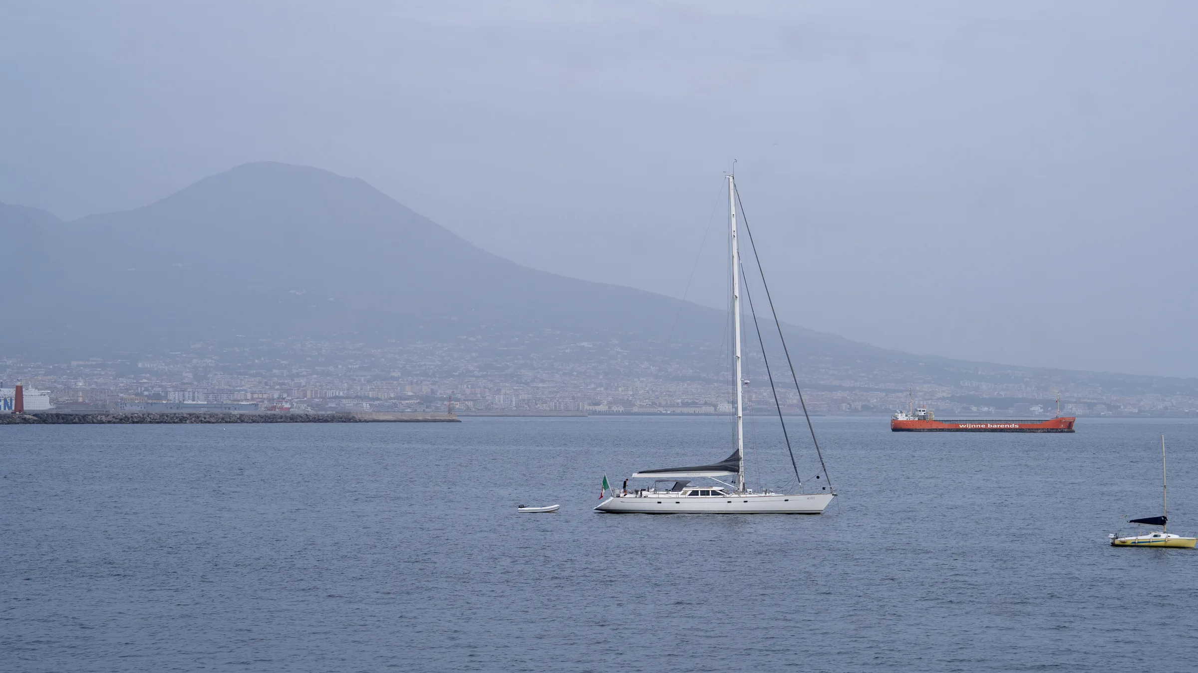 A sailing yacht anchored in the Gulf of Naples with Mount Vesuvius rising hazy behind it.