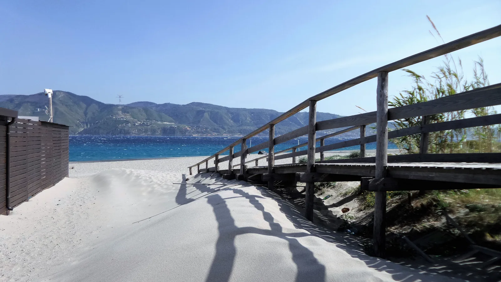 A wooden boardwalk crossing white dunes toward the Strait of Messina at Capo Peloro.