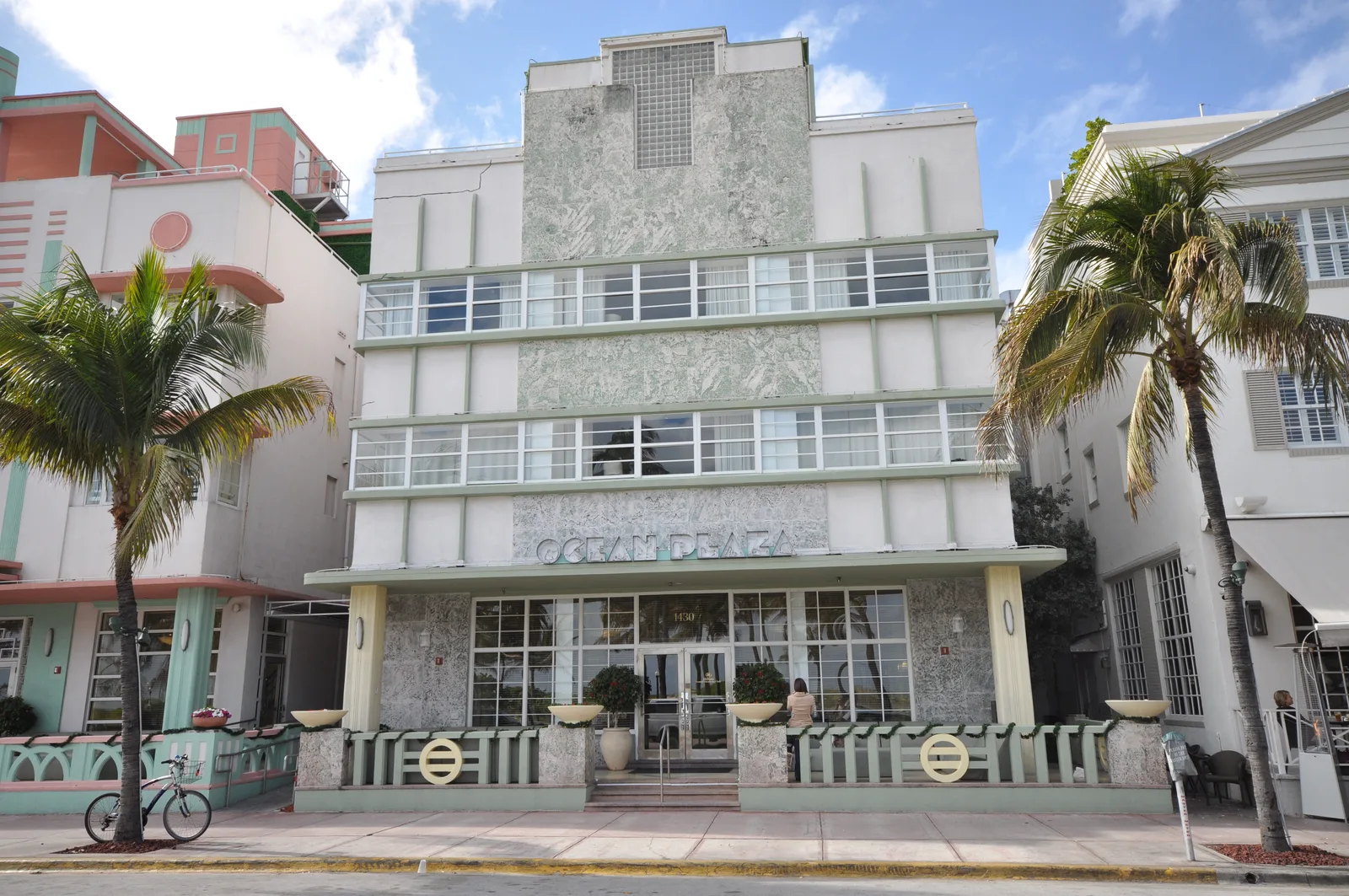 A symmetrical white and pastel Art Deco hotel facade on Ocean Drive with palm trees and a parked bicycle.