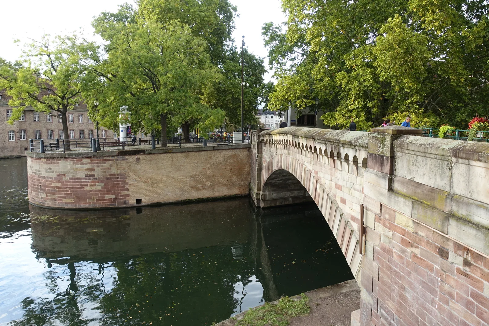 A stone arch bridge over a river arm in Strasbourg with trees on the bank and a Ponts Couverts tower beyond.