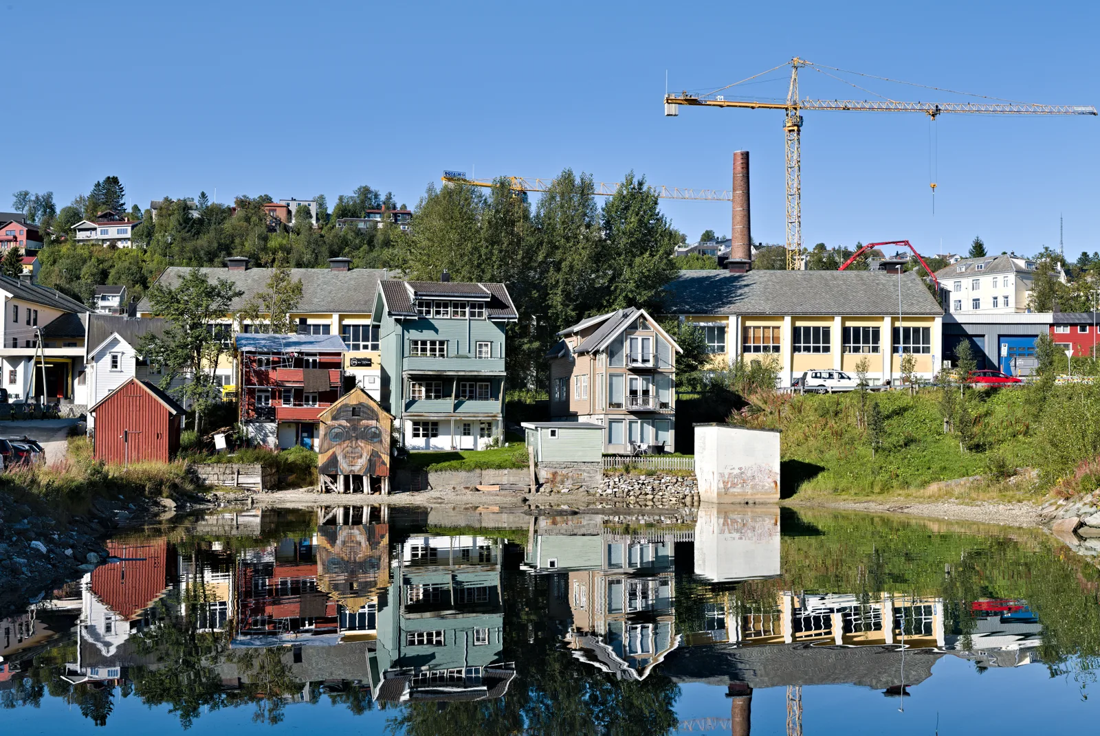 Tromso houses and boathouses reflected in a still bay at Juldagan under a blue sky with a construction crane behind.