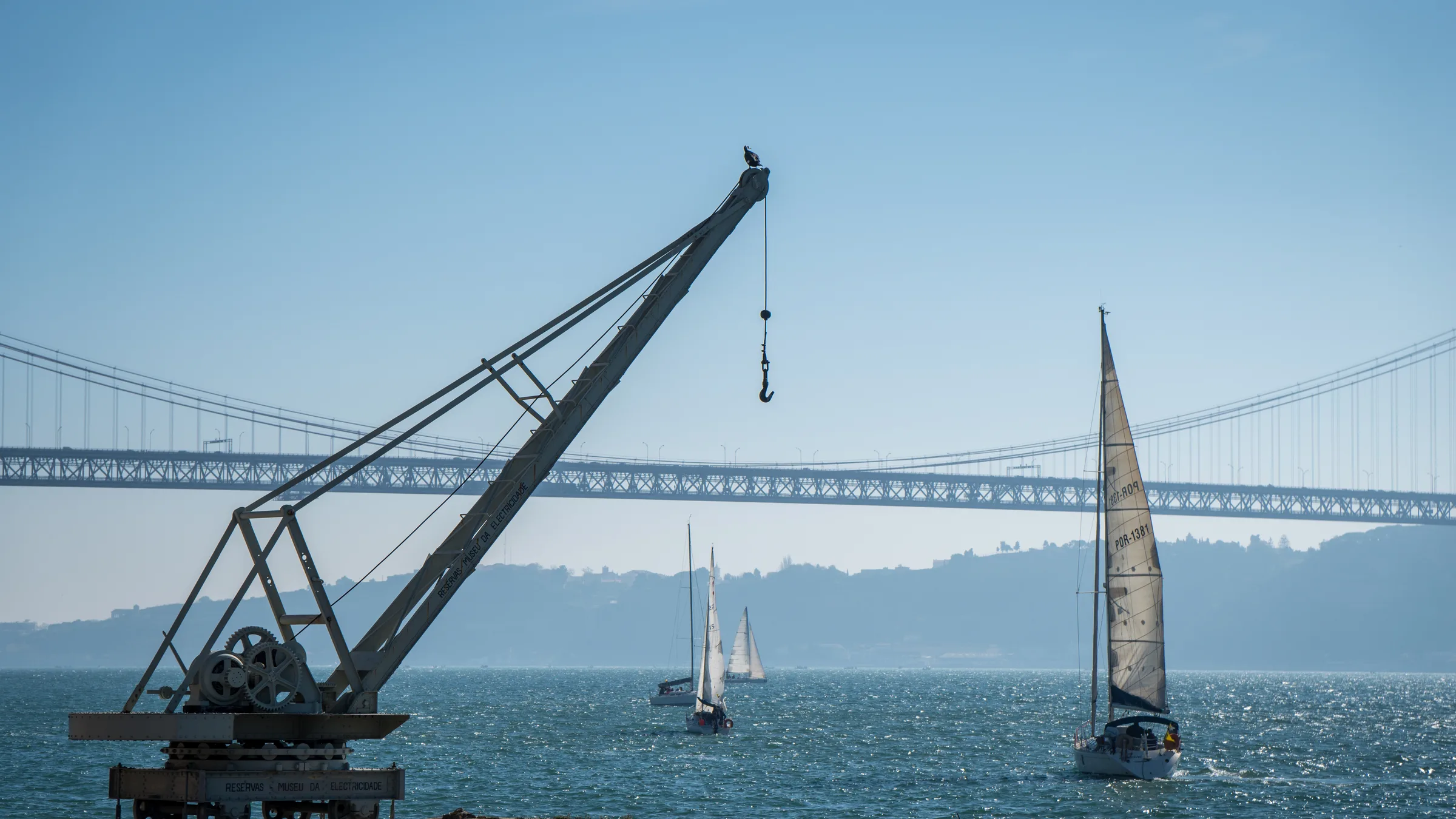 A vintage dockside crane on the Tagus with the 25 de Abril Bridge and sailing boats in the background.