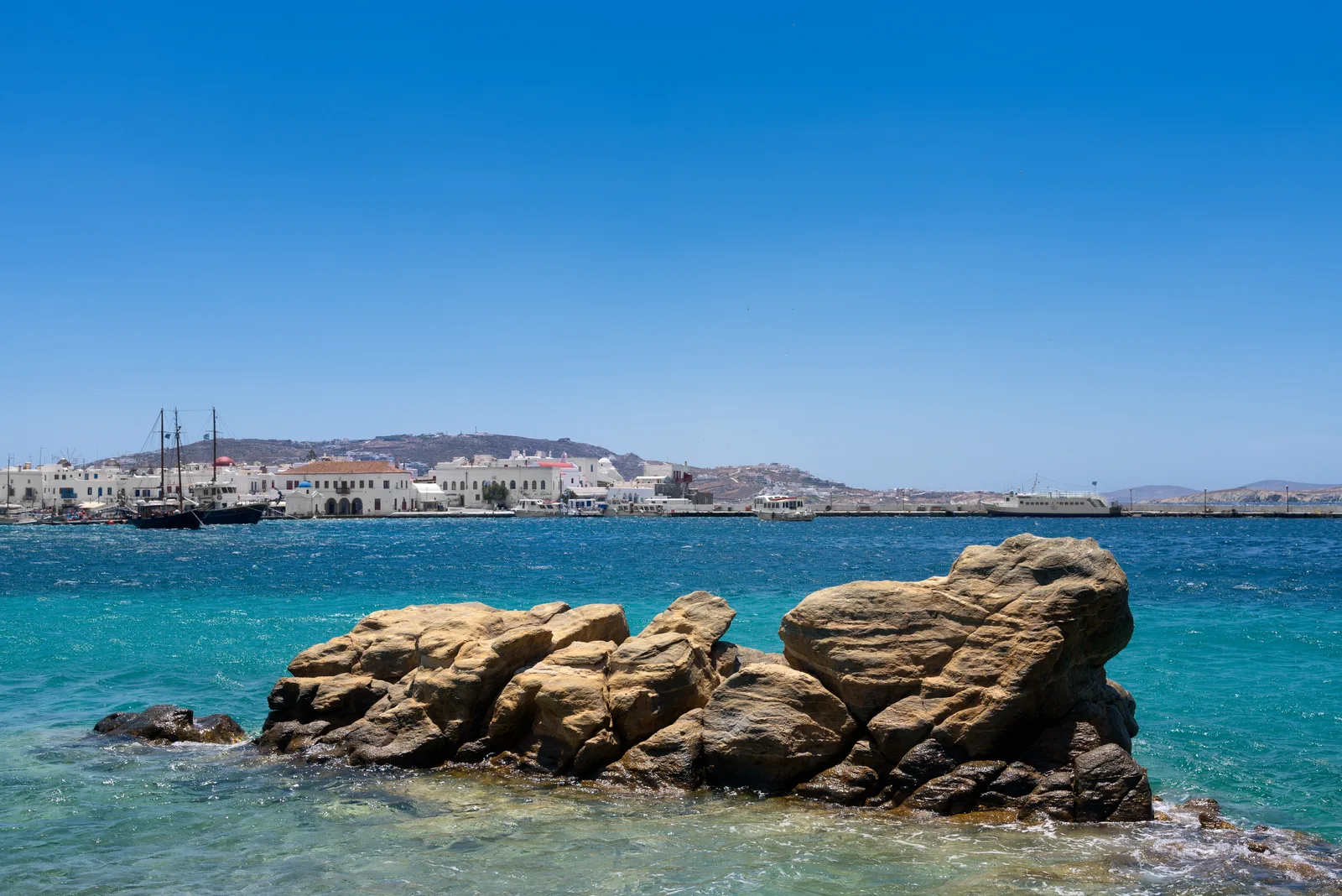 A tumble of sunlit rocks in Mykonos harbour with a tall ship and the town on the horizon.