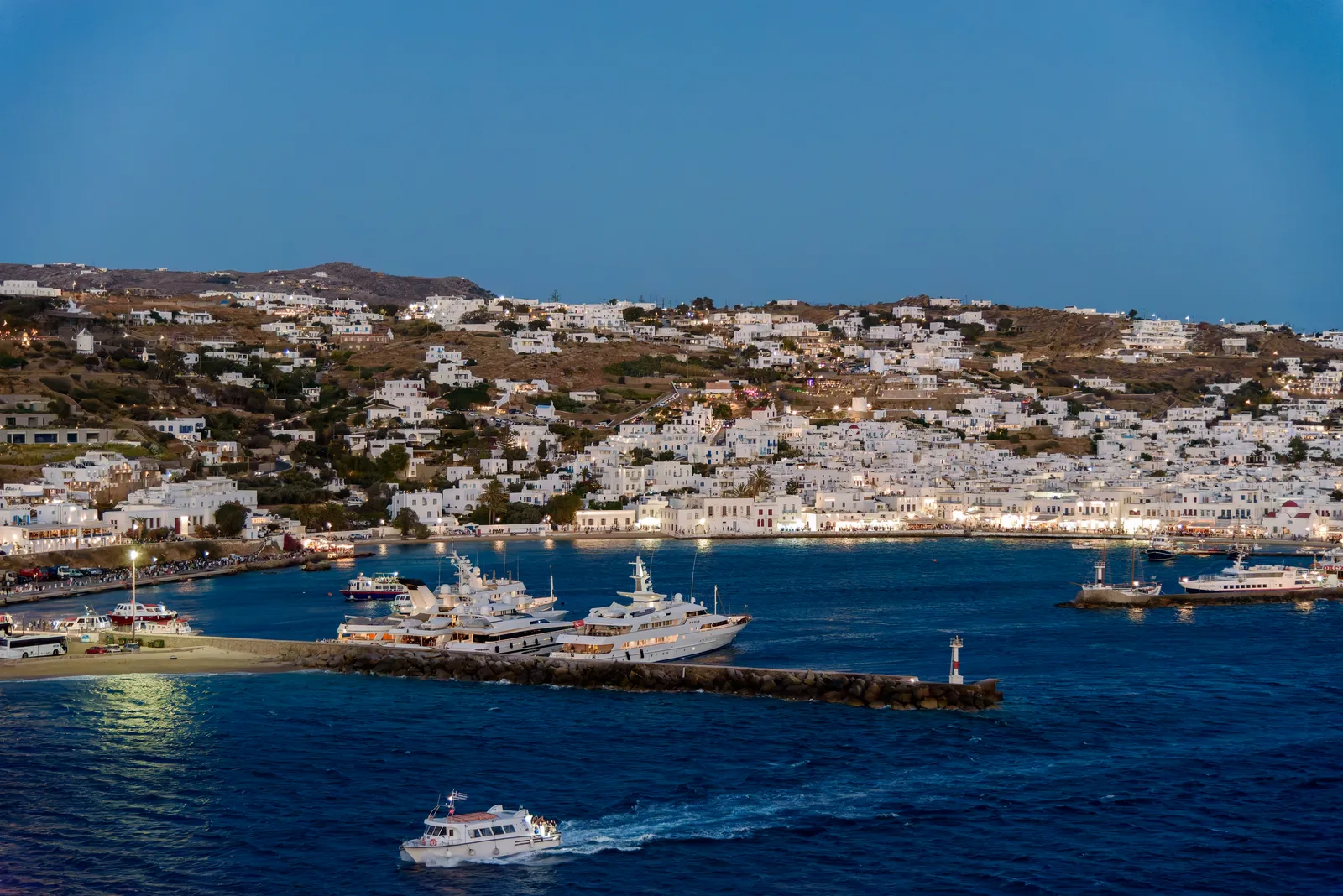 Mykonos harbour at dusk with yachts and ferries by the breakwater and the lit white town above.