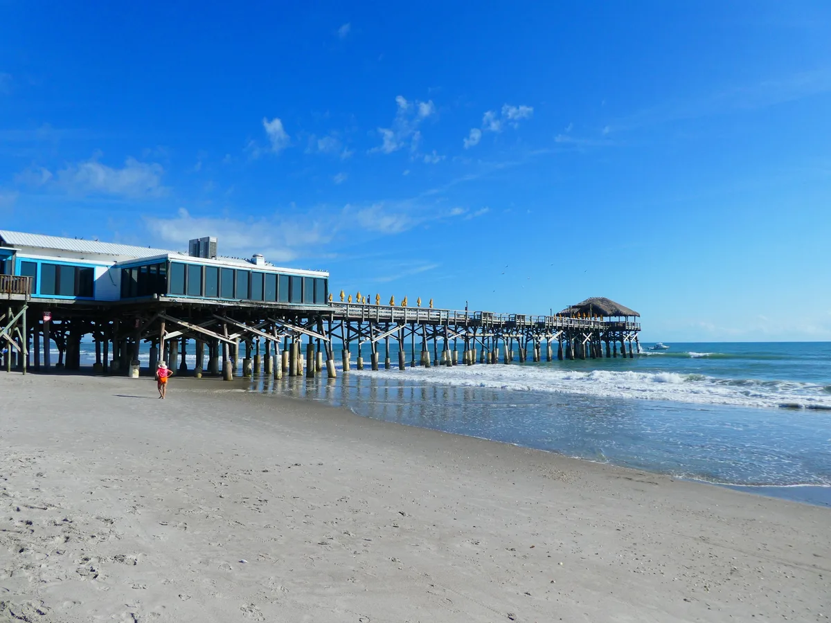 Cocoa Beach Pier stretching out over the surf from a wide sandy beach on a bright blue day.