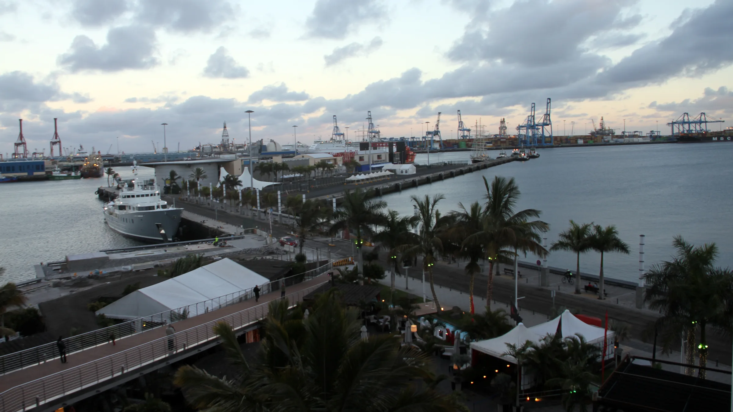 The Las Palmas port waterfront with palm-lined promenade, a moored motor yacht and the container cranes of the commercial harbour in the distance.