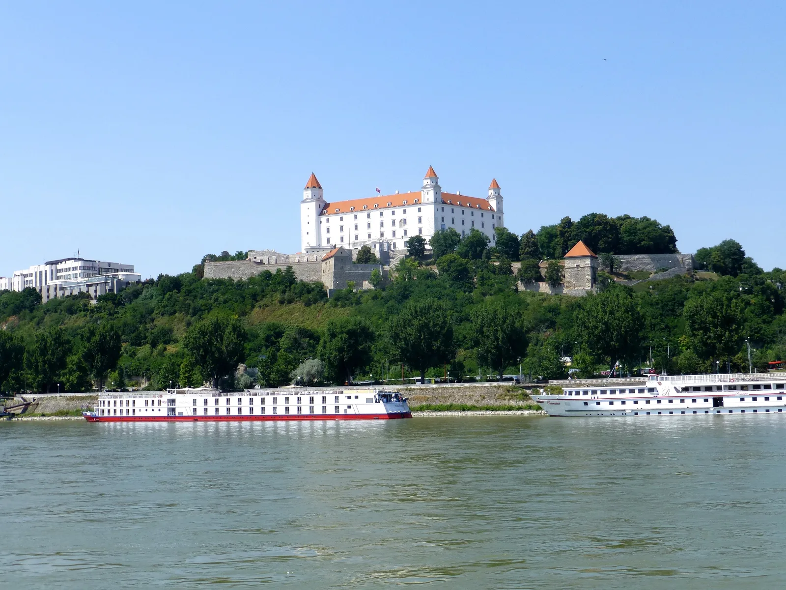 Bratislava Castle on its green hill above the Danube with two white river ships moored at the bank.