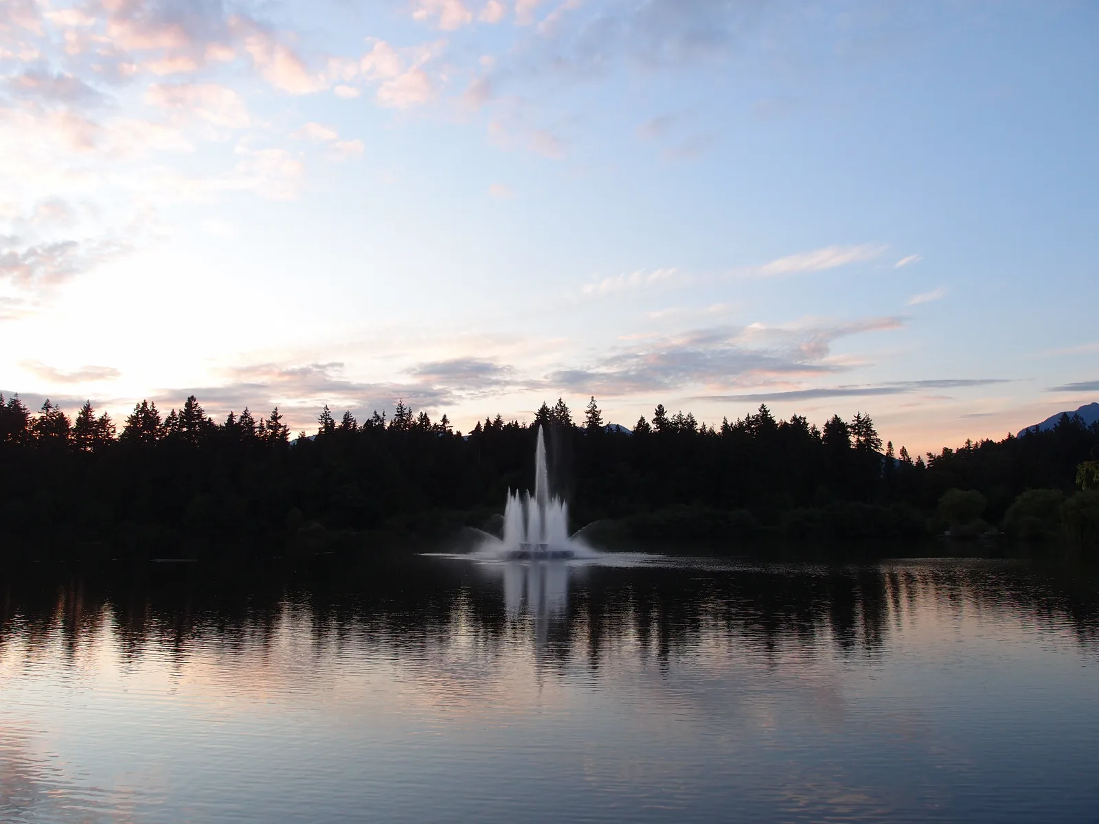 A fountain in Lost Lagoon at dusk beneath a pink and blue sky, with trees reflected in the still water.