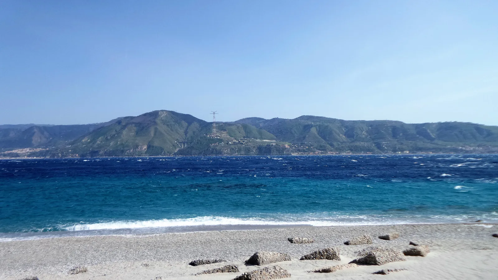 A sandy shore and clear water at Capo Peloro on the Strait of Messina, with the Calabrian hills across the strait.