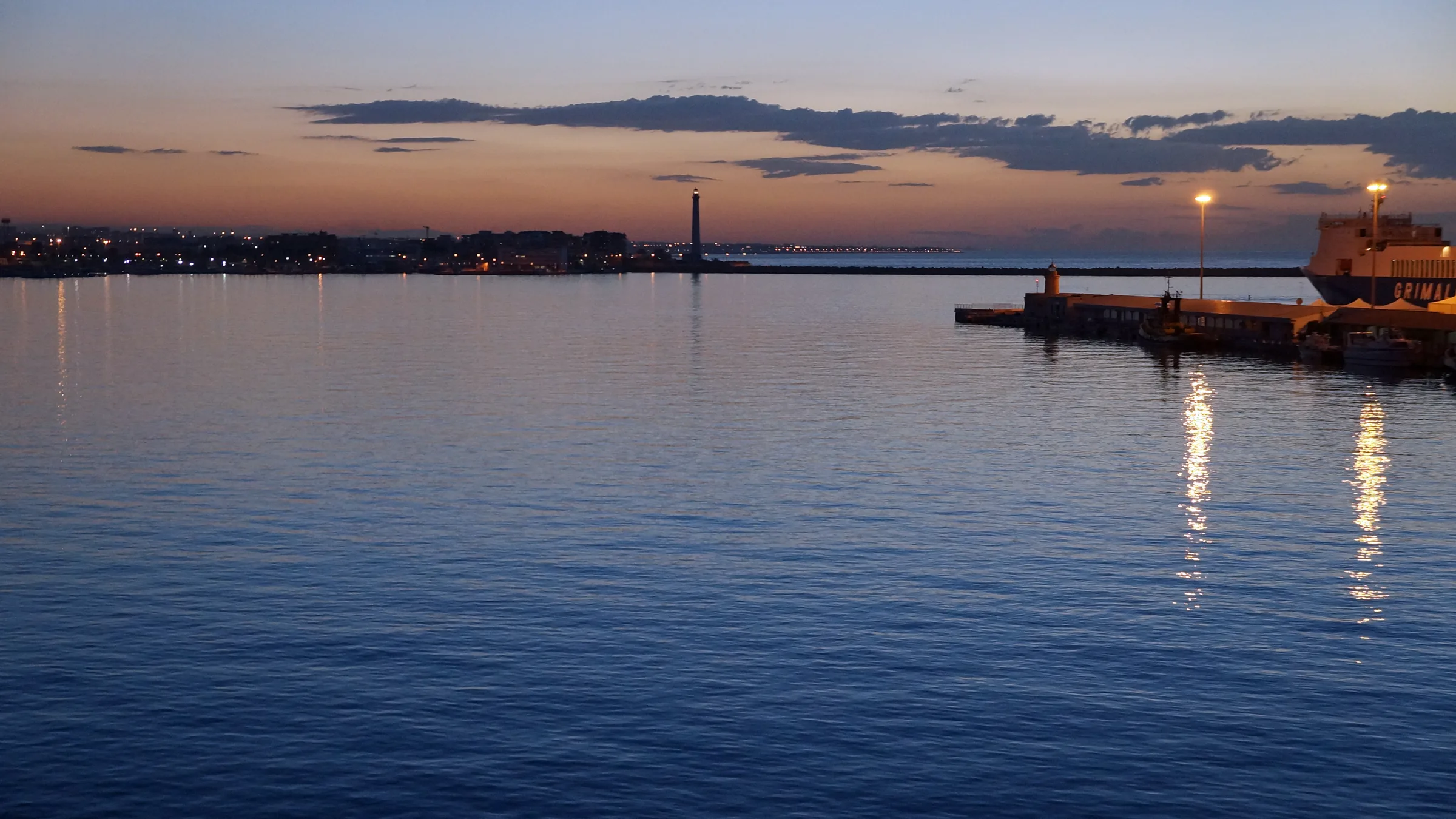 Dusk over the calm waters of Bari harbour with a lighthouse and quay lights on the horizon.