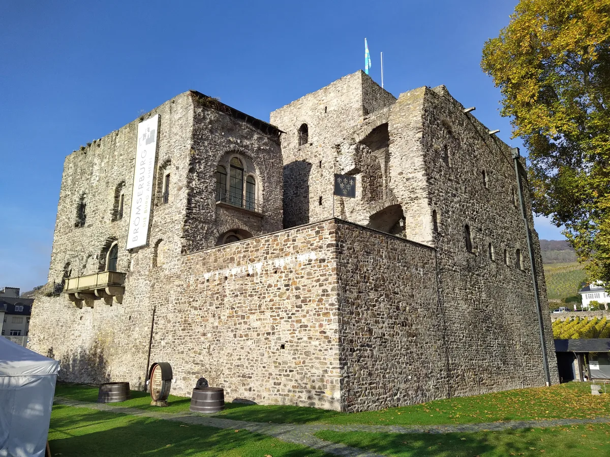 The stone Brömserburg castle on the Rhine bank at Rüdesheim with a green lawn and wine barrels in front.