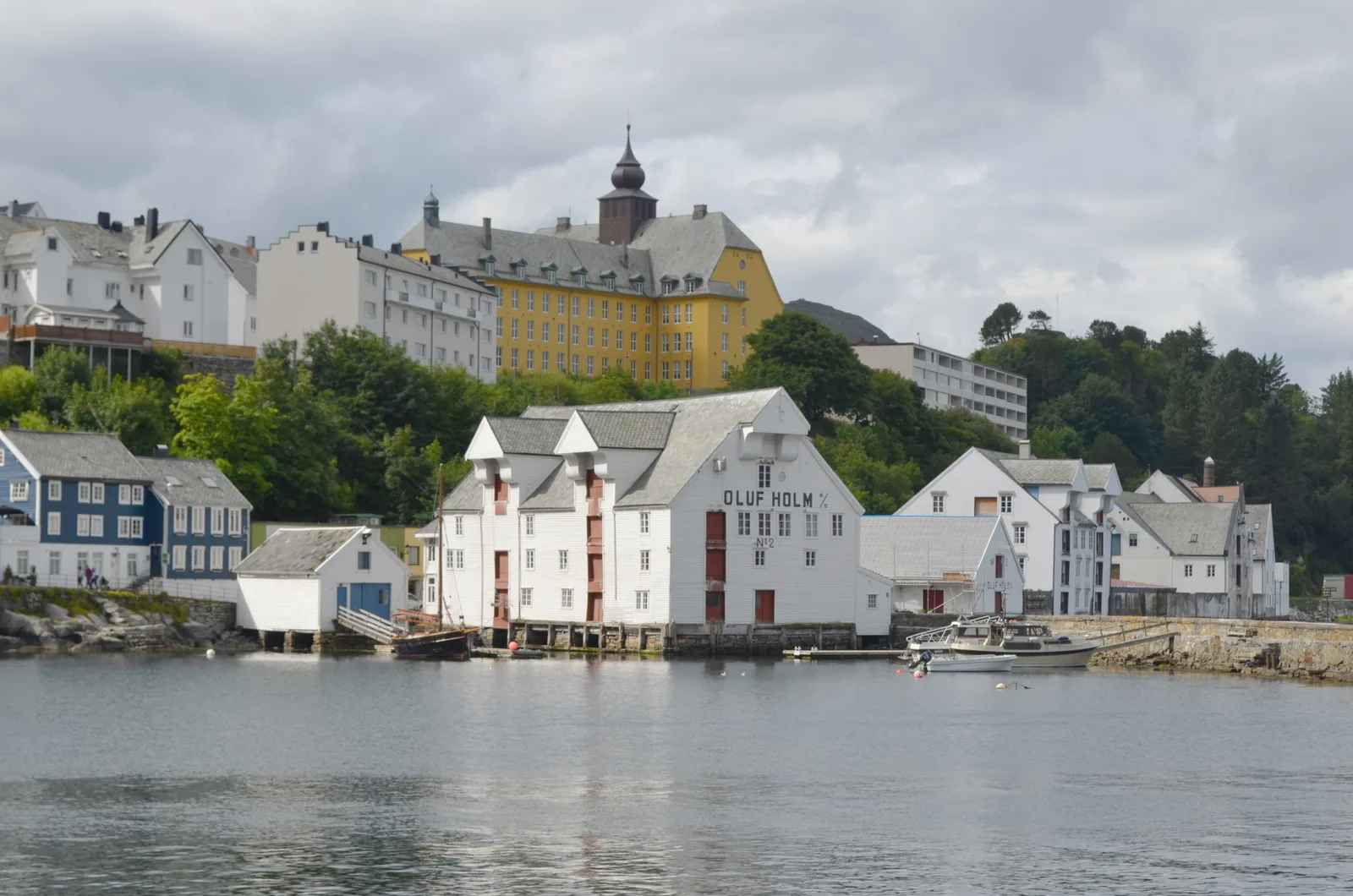 Whitewashed timber warehouses and an old hilltop school building on Alesund's quayside, with moored boats.