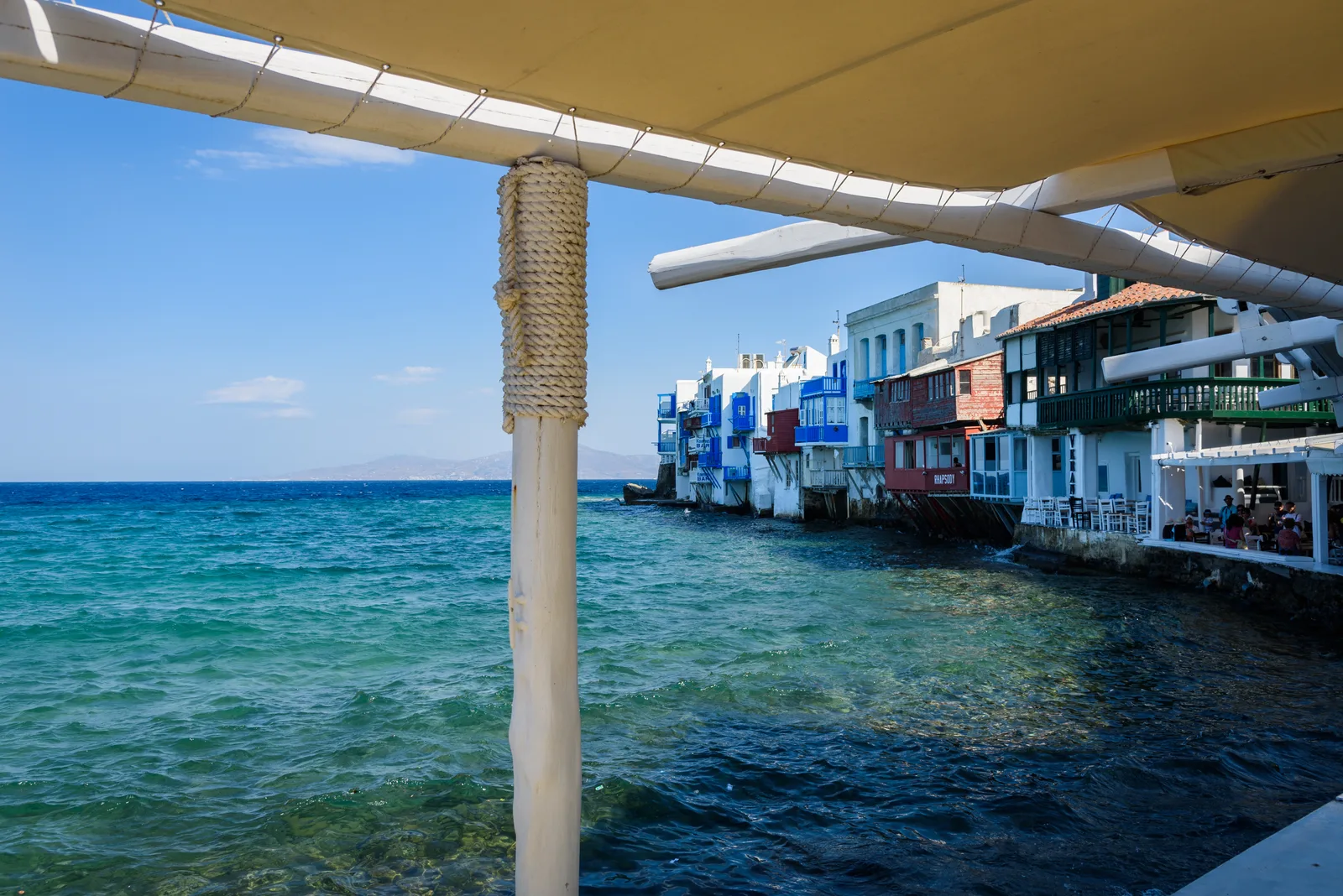 White and coloured balconied houses at the water's edge of Little Venice in Mykonos seen from under a cafe awning, with a deep blue sea.