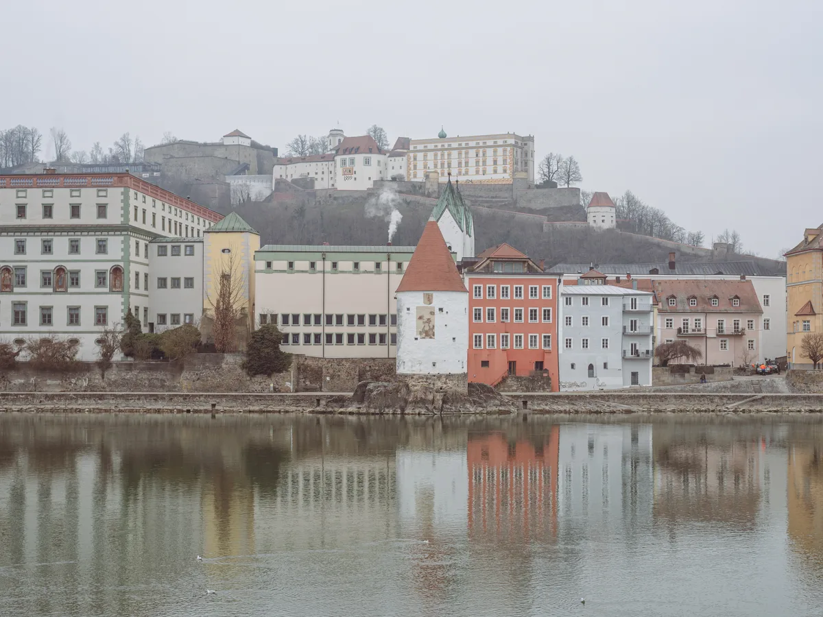 The Inn riverfront in Passau with the Schaiblingsturm tower and old-town buildings reflected in the water.