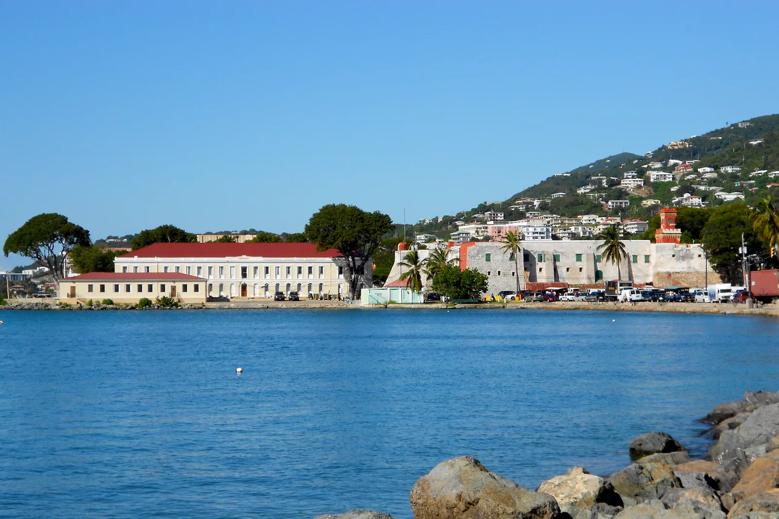 The Charlotte Amalie waterfront with a low red-roofed building, green hillside and rocks in the foreground.