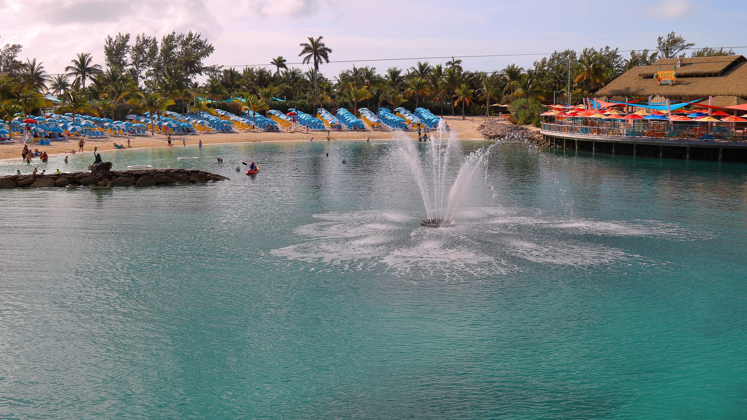 A calm lagoon with a fountain, rows of blue sun loungers and a thatched building across the water at CocoCay.