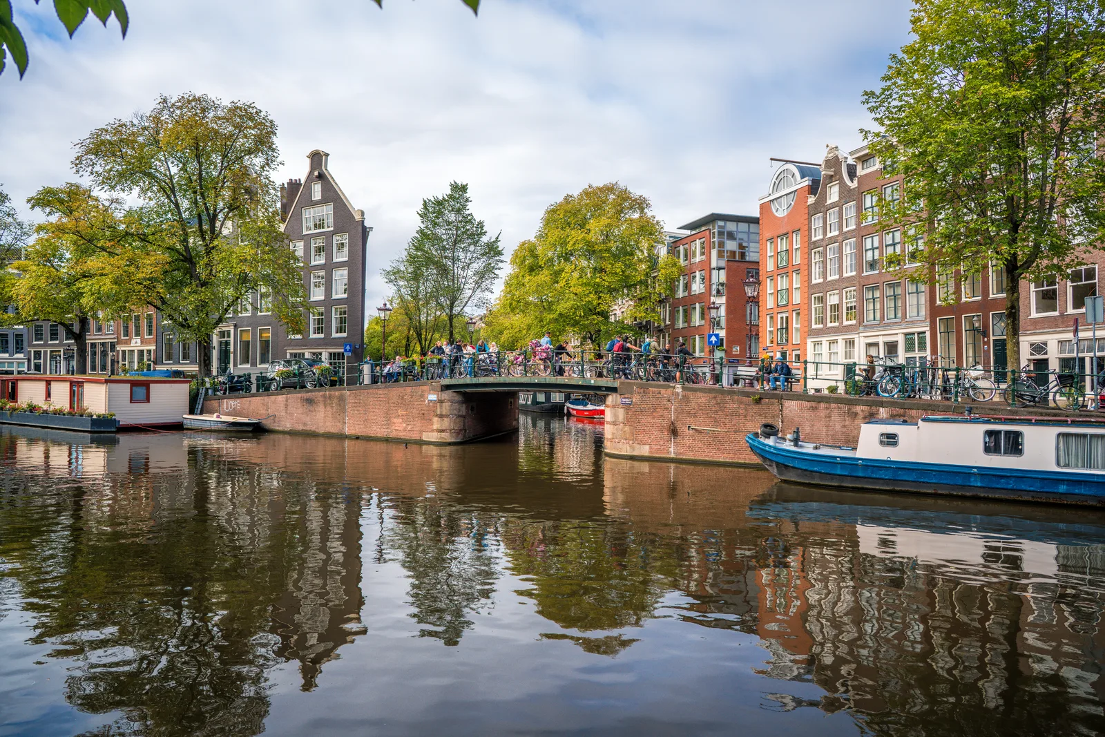 A canal junction in Amsterdam with a brick bridge, reflected autumn trees and a blue houseboat.