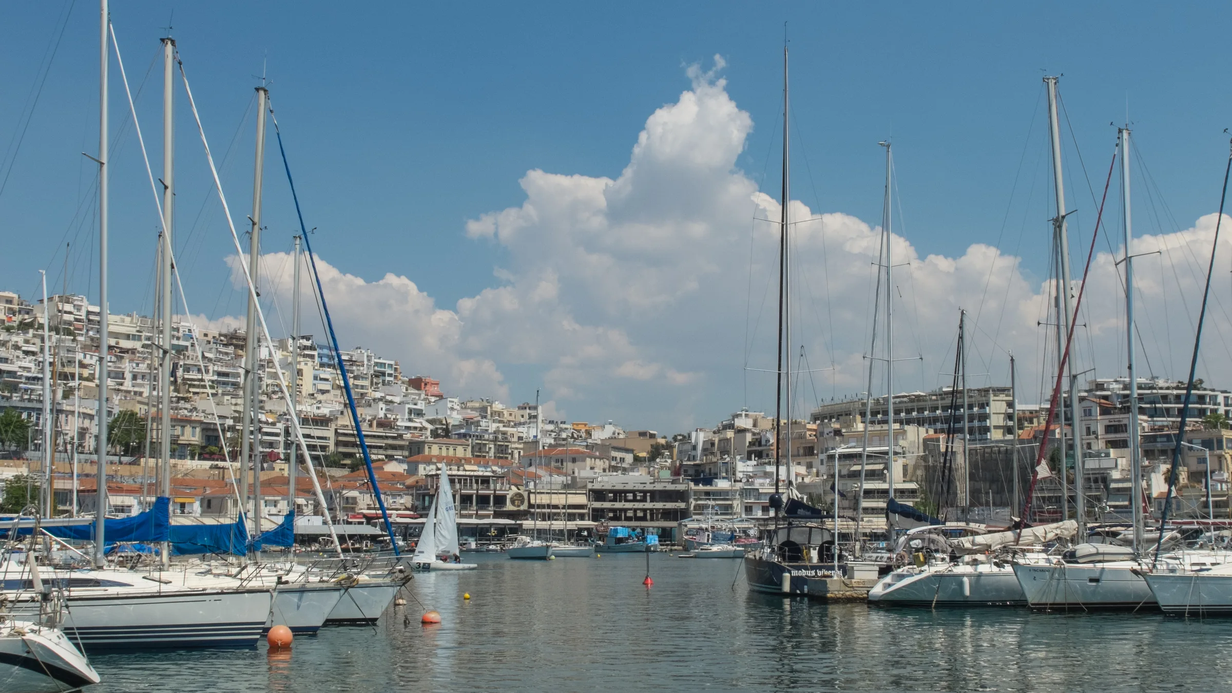 Yachts moored at the Mikrolimano marina in Piraeus with a big cumulus cloud and white apartment blocks behind.